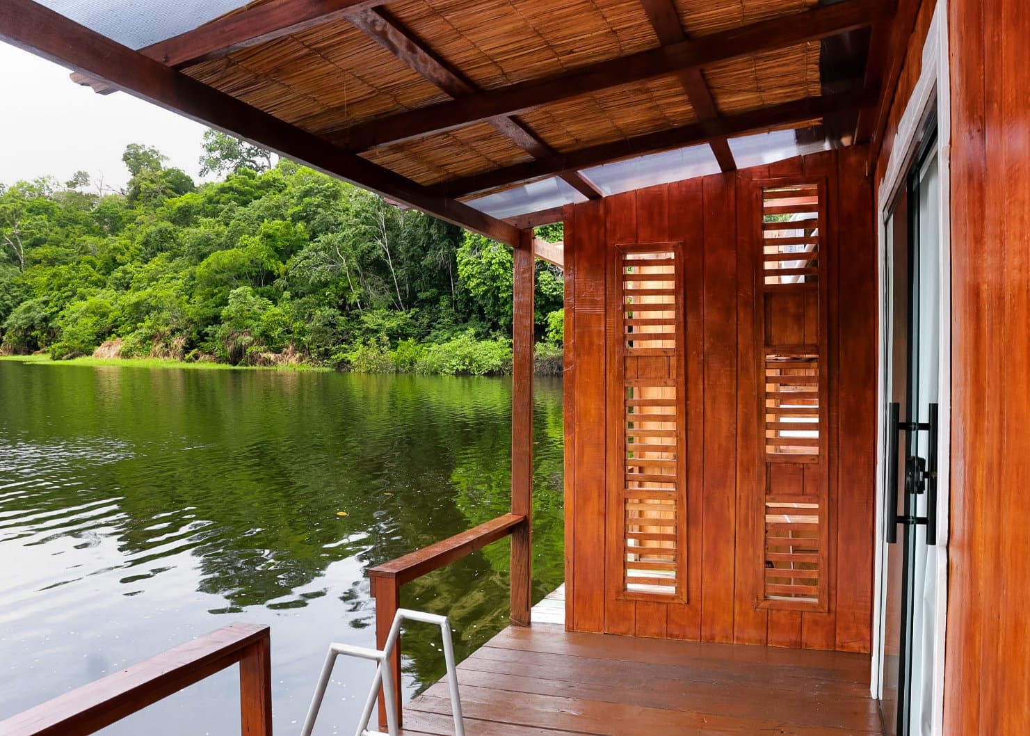 Madeira, água escura e Amazônia em cada direção. Deck de madeira nobre e varanda com telhado de palha de lodge ecológico em tour pela selva amazônica brasileira, com venezianas de madeira voltadas para um rio de águas pretas e floresta densa ao fundo