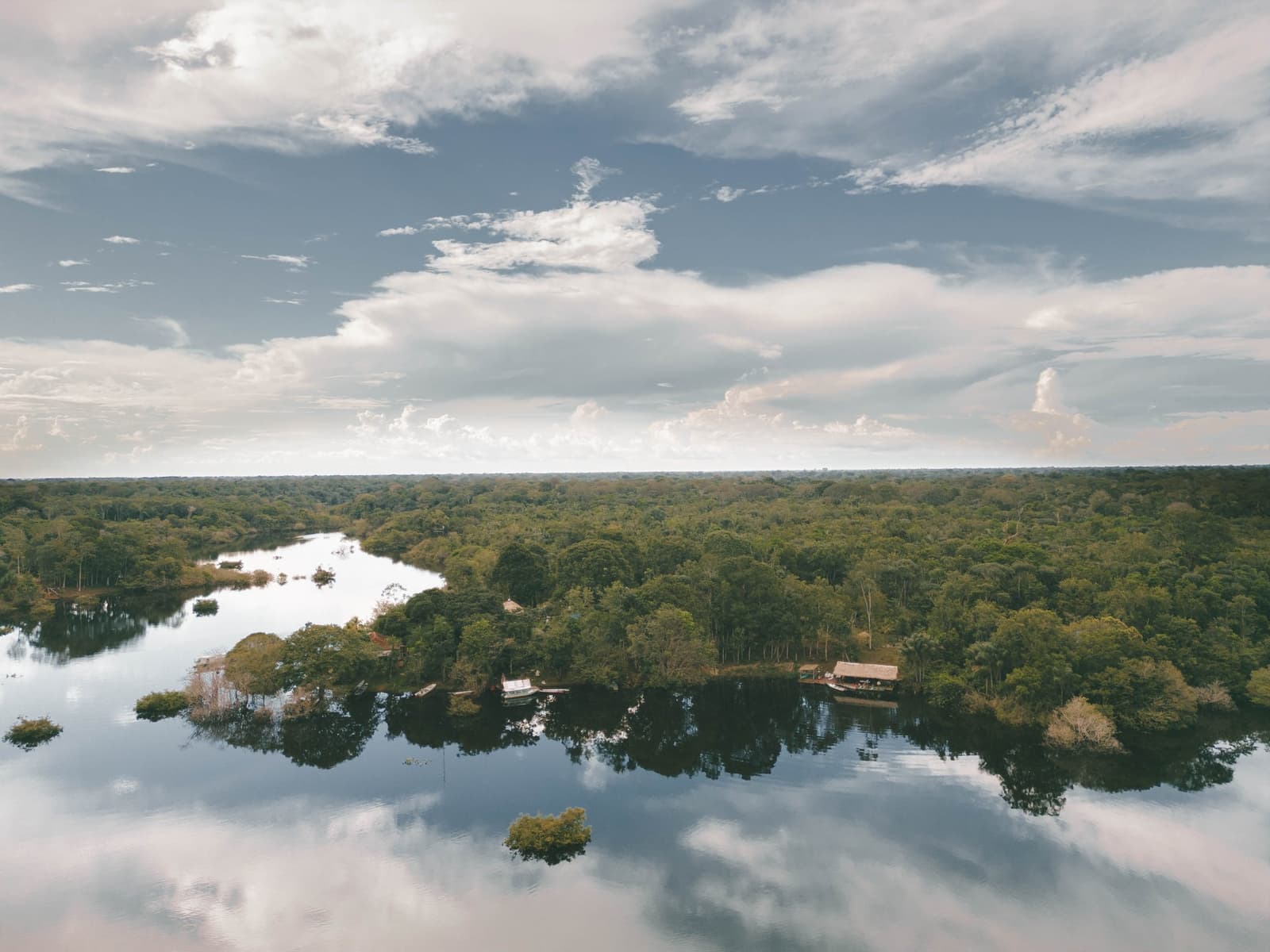 Copa das árvores até o horizonte, o lodge quieto à beira d'água. Vista aérea de um rio de águas escuras e calmas serpenteando pela floresta tropical densa em um tour pela selva amazônica brasileira, com estruturas de lodge com telhado de palha e um pequeno barco visíveis à beira da margem arborizada