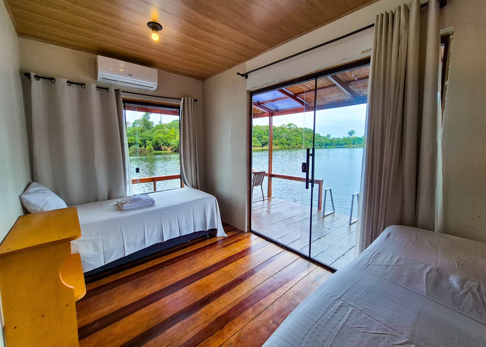 Conforto à beira do rio, com a floresta a poucos metros. Quarto duplo em lodge na Amazônia com piso de madeira polida, ar-condicionado e porta de vidro deslizante abrindo para uma varanda coberta com vista para um rio tranquilo cercado de floresta densa