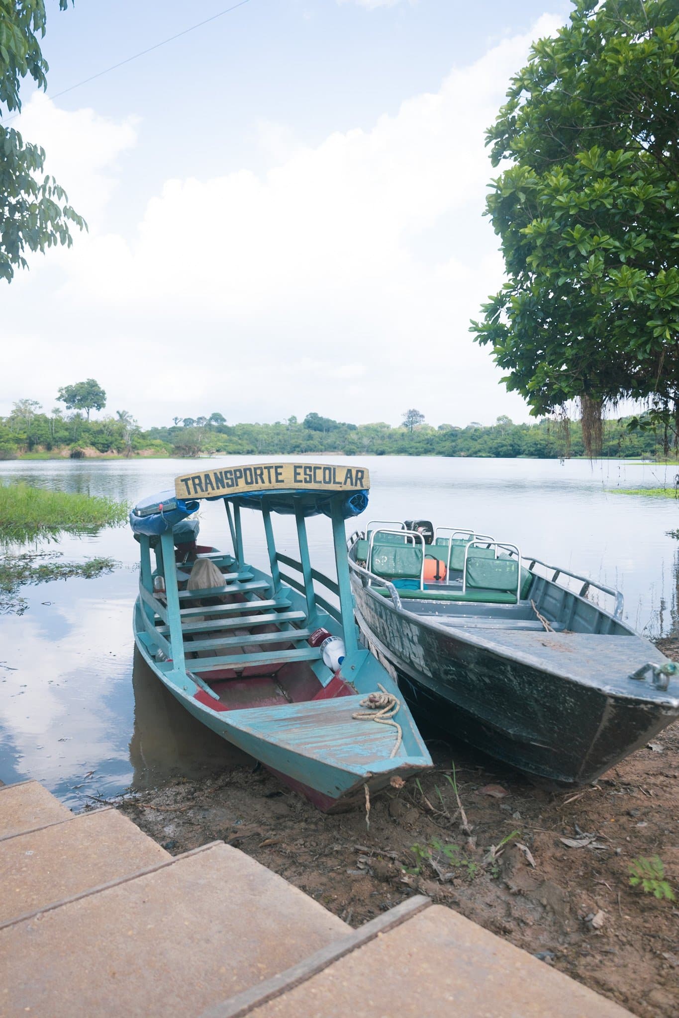 Ribeirinhos chegam de barco — a Amazônia tem seus próprios caminhos. Uma embarcação de madeira turquesa com a inscrição 'Transporte Escolar' e um segundo barco a motor atracados numa margem de terra batida, com o rio calmo e vegetação tropical densa ao fundo