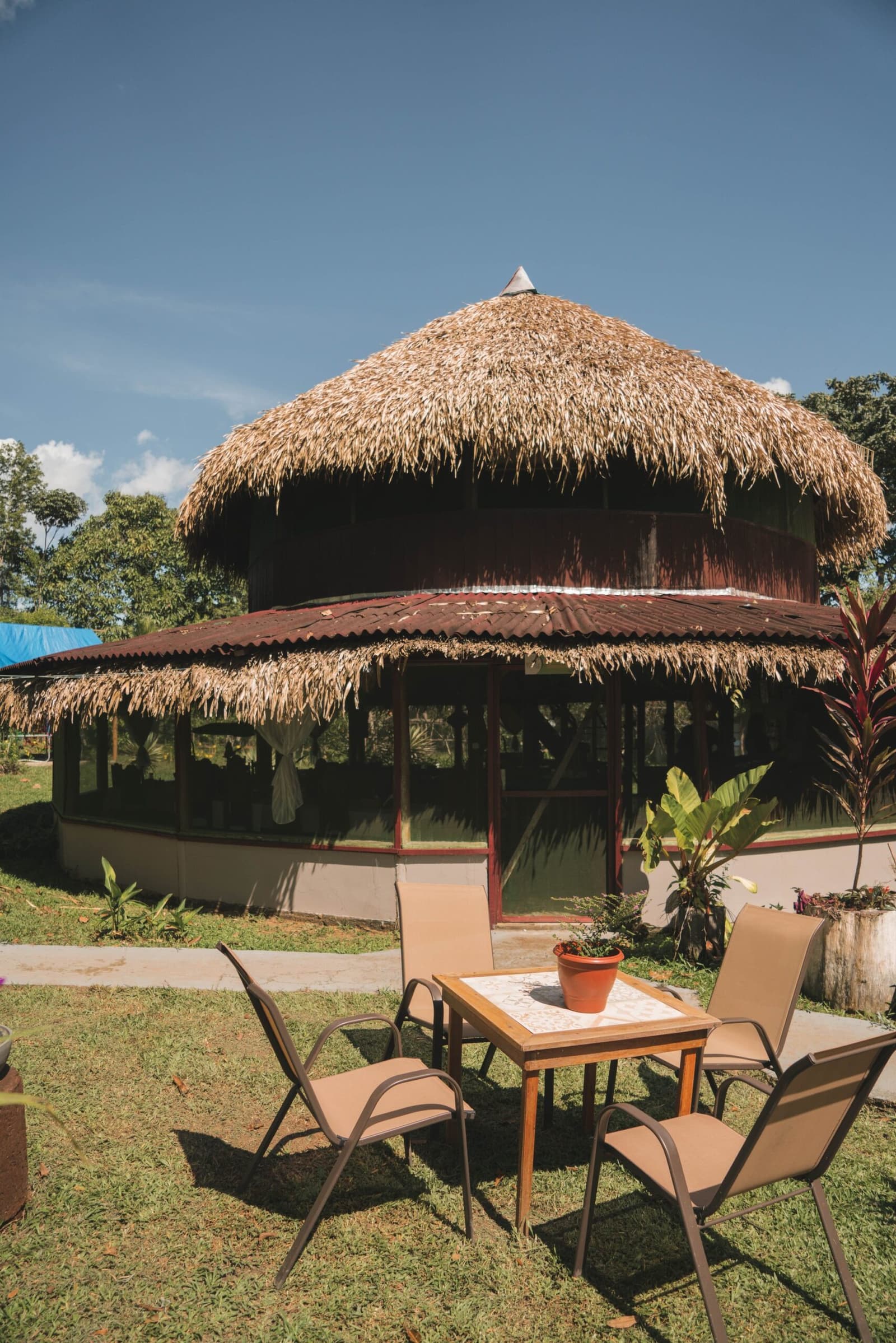 Redondo, coberto de palha e aberto para a floresta — seu refúgio na Amazônia. Photo: Yuri Panichi Um lodge circular de dois andares com telhado de palha rodeado por vegetação tropical em um tour pela selva amazônica brasileira, com mesa e cadeiras em gramado ensolarado no primeiro plano