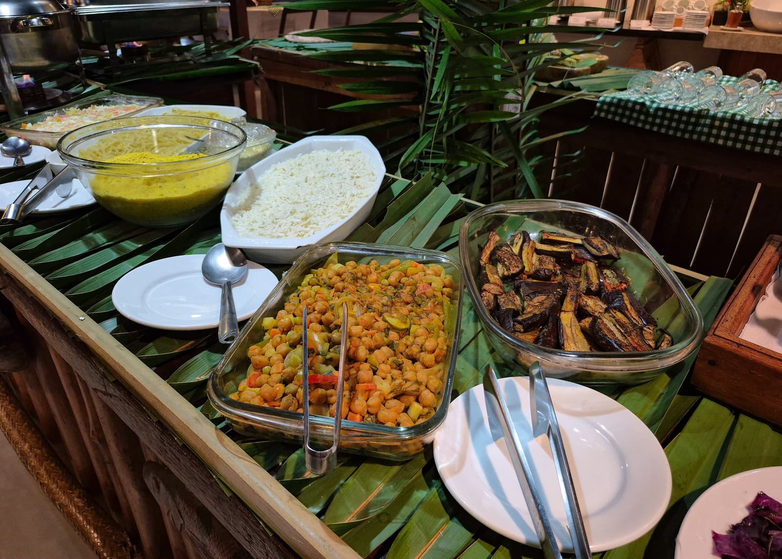 Pratos fartos e naturais para repor energia antes de mais uma jornada pela floresta. Um colorido buffet vegetariano em mesa decorada com folhas de palmeira, com grão-de-bico temperado, berinjela assada, arroz branco e molho de curry amarelo