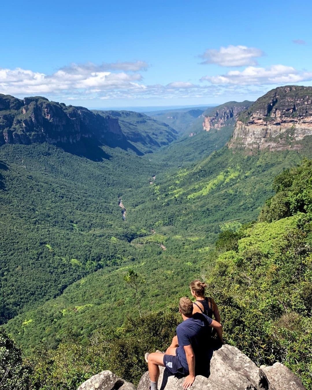 Quand la vallée parle mieux que tous les mots. Deux randonneurs assis sur un rocher surplombant un canyon boisé lors d'une randonnée Chapada Diamantina, avec des falaises de grès et une rivière serpentant loin en contrebas
