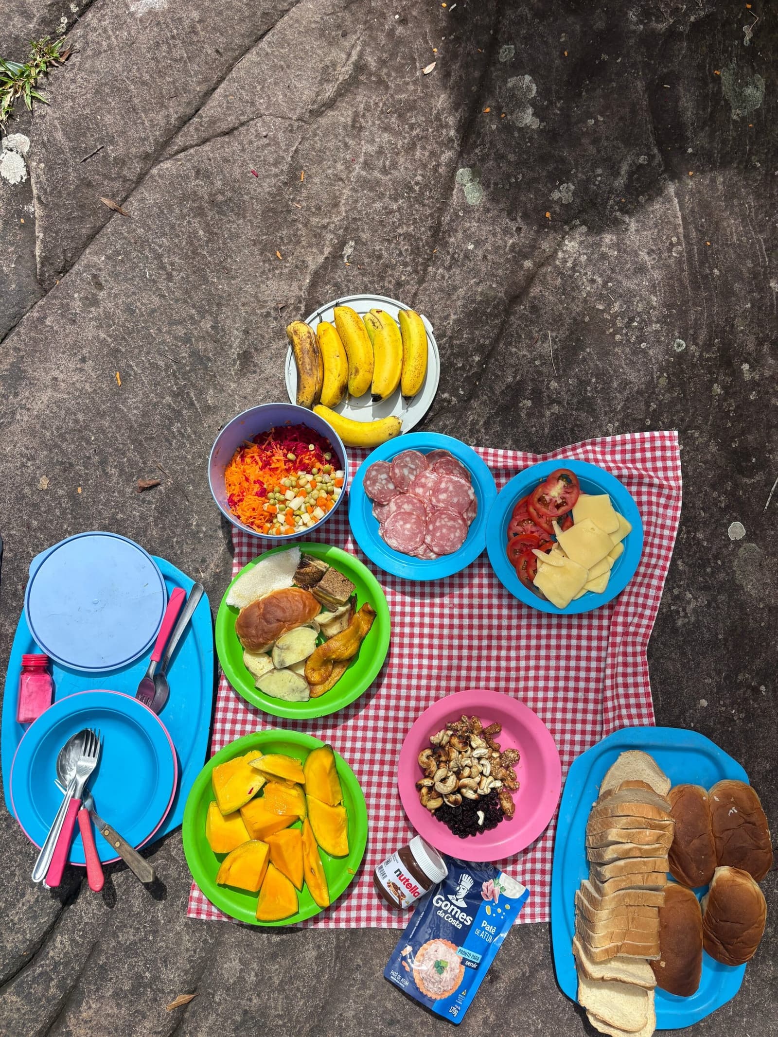 Le meilleur repas du trek, servi sur granit. Vue de dessus d'un pique-nique coloré disposé sur un rocher plat lors d'une randonnée dans la Chapada Diamantina, avec des assiettes de fruits, salami, fromage, tomates, légumes râpés, noix de cajou, raisins secs, bananes, sandwiches et pain tranché sur une nappe à carreaux rouges