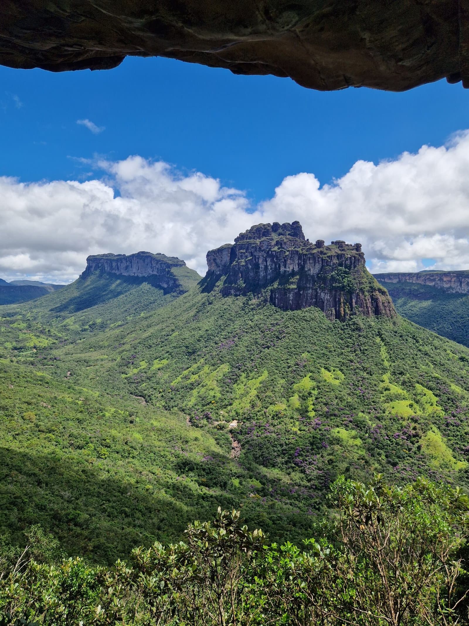 Le rocher fait le cadrage, il ne reste qu'à marcher. Un surplomb rocheux encadre la vue sur un mesa aux parois verticales s'élevant au-dessus de collines verdoyantes lors d'une randonnée dans la Chapada Diamantina, sous un ciel bleu vif ponctué de nuages blancs