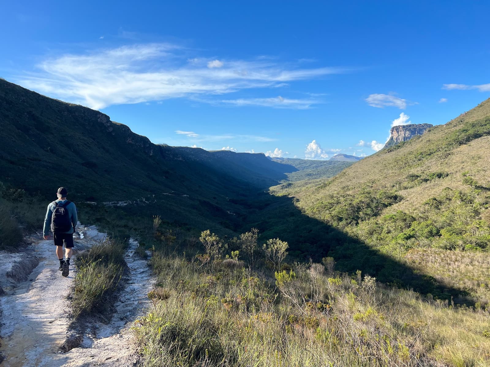 Un sentier, une vallée immense, et le silence qui fait du bien. Un randonneur avec un sac à dos marche sur un sentier de crête rocailleux lors d'une randonnée à la Chapada Diamantina, avec une vaste vallée verdoyante encadrée d'escarpements gréseux sous un ciel bleu