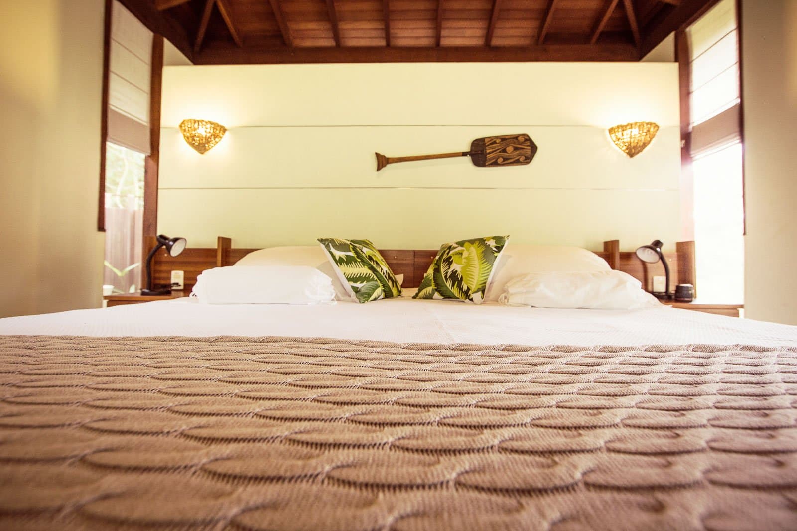 Strak opgemaakt bed, een houten peddel aan de muur, en het oerwoud net om de hoek. Kingsize bed met tropische bladmotiefkussens, houten hoofdbord, gevlochten wandlampen en een gesneden peddel aan de muur in een bungalow van Cristalino Jungle Lodge