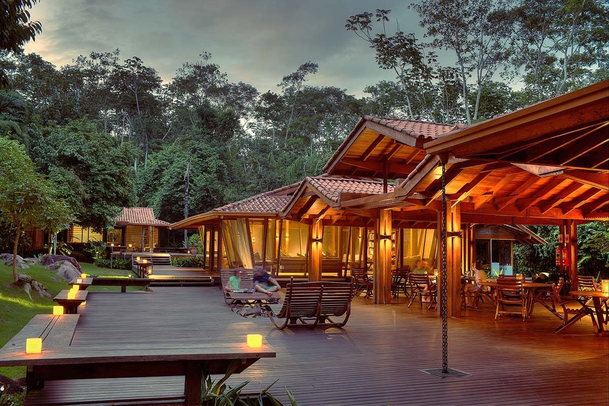 Dinner hour, and the forest is already leaning in. Cristalino Jungle Lodge's timber open-air deck glowing with candles and warm lamp light at dusk, with guests relaxing on wooden rocking chairs and dense rainforest rising in the background