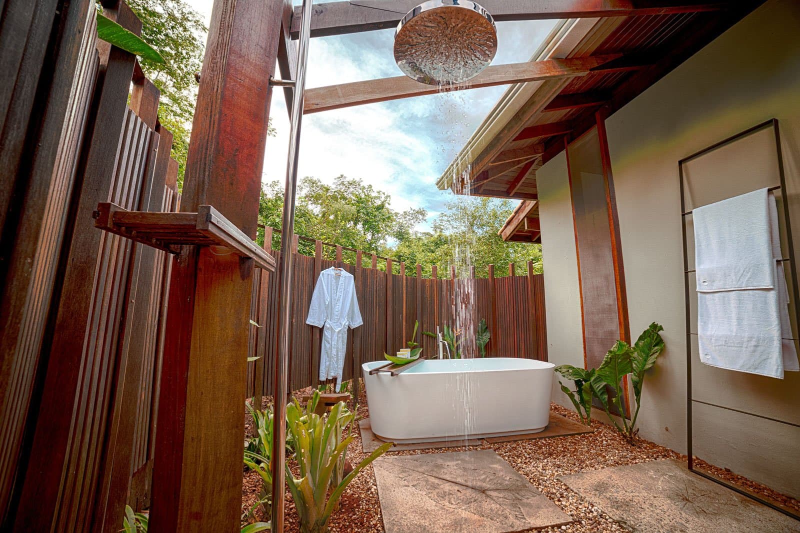 Baden onder de Amazone-hemel, met een badjas die al op je wacht. Een open badkamer van Cristalino Jungle Lodge met een vrijstaand wit ligbad onder een regendouche, omgeven door tropische planten, donkere houten schermen en het bladerdak van het regenwoud