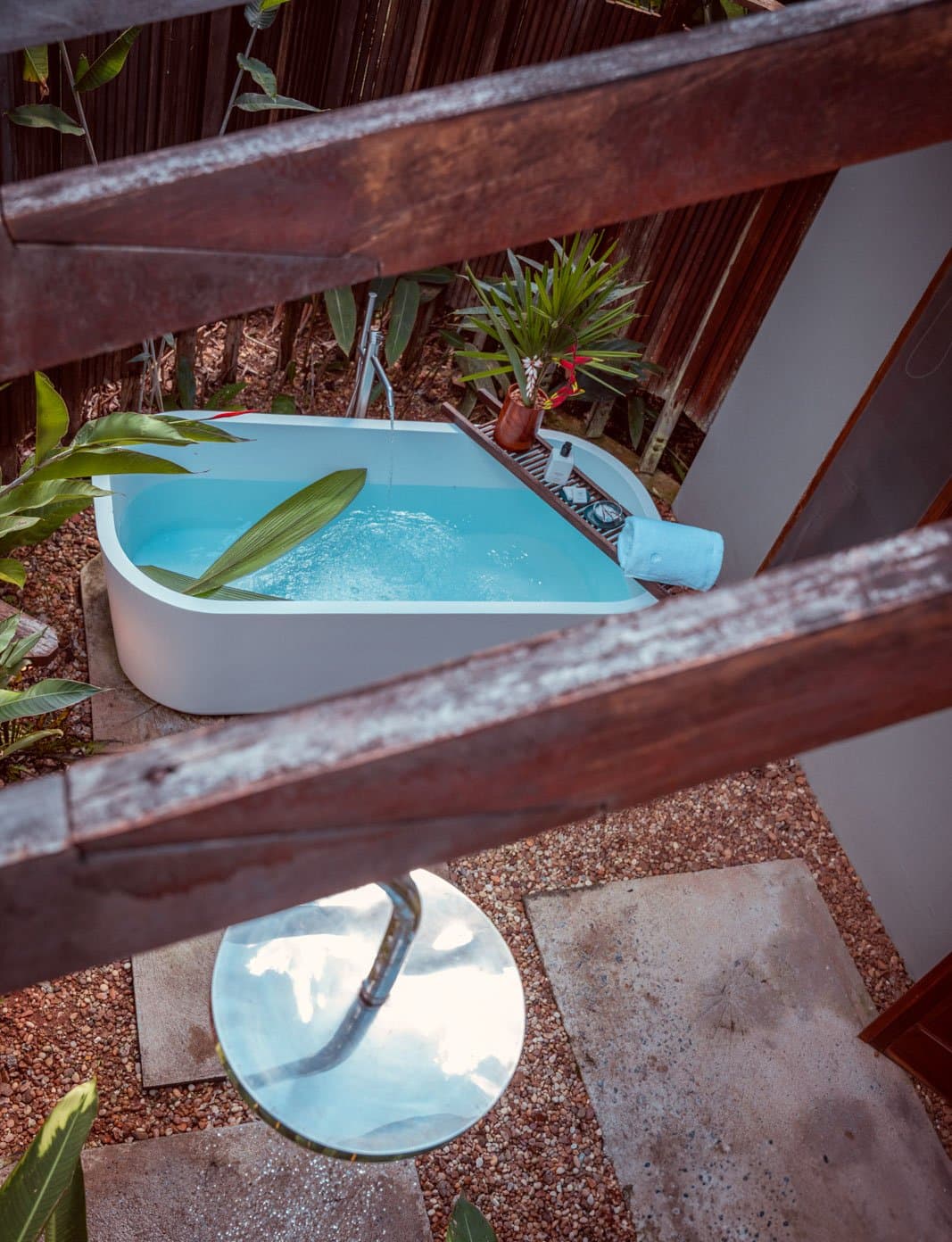 Inweekten in de jungle, terwijl het oerwoud op je wacht. Een vrijstaand bubbelbad in de openlucht gevuld met helder blauw water bij Cristalino Jungle Lodge, omgeven door tropisch gebladerte, een houten badplank met bloemen en toiletartikelen, en een ronde buitendouche eronder, van bovenaf gefotografeerd door een rustieke houten reling