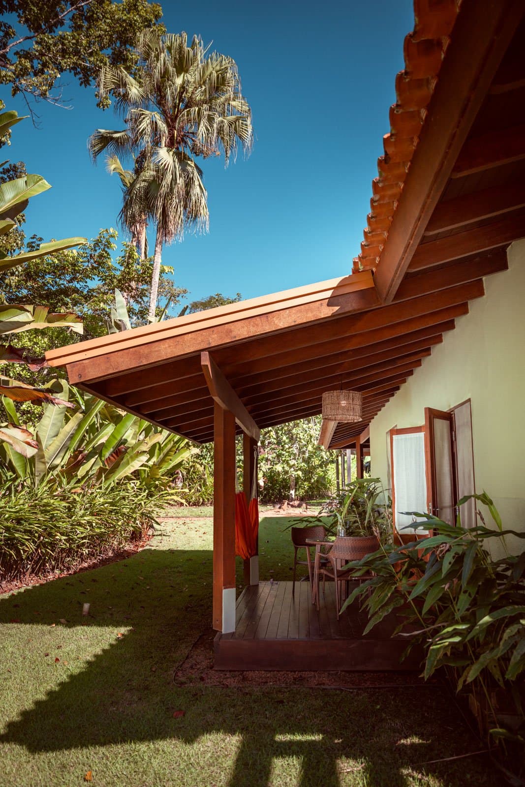 Koele schaduw, een rieten lamp en het regenwoud op een steenworp afstand. Beschutte hardhouten veranda van Cristalino Jungle Lodge met een rotan hanglamp, rotan stoelen en een weelderige tropische tuin met bananenplanten en een hoge palm onder een diepblauwe hemel
