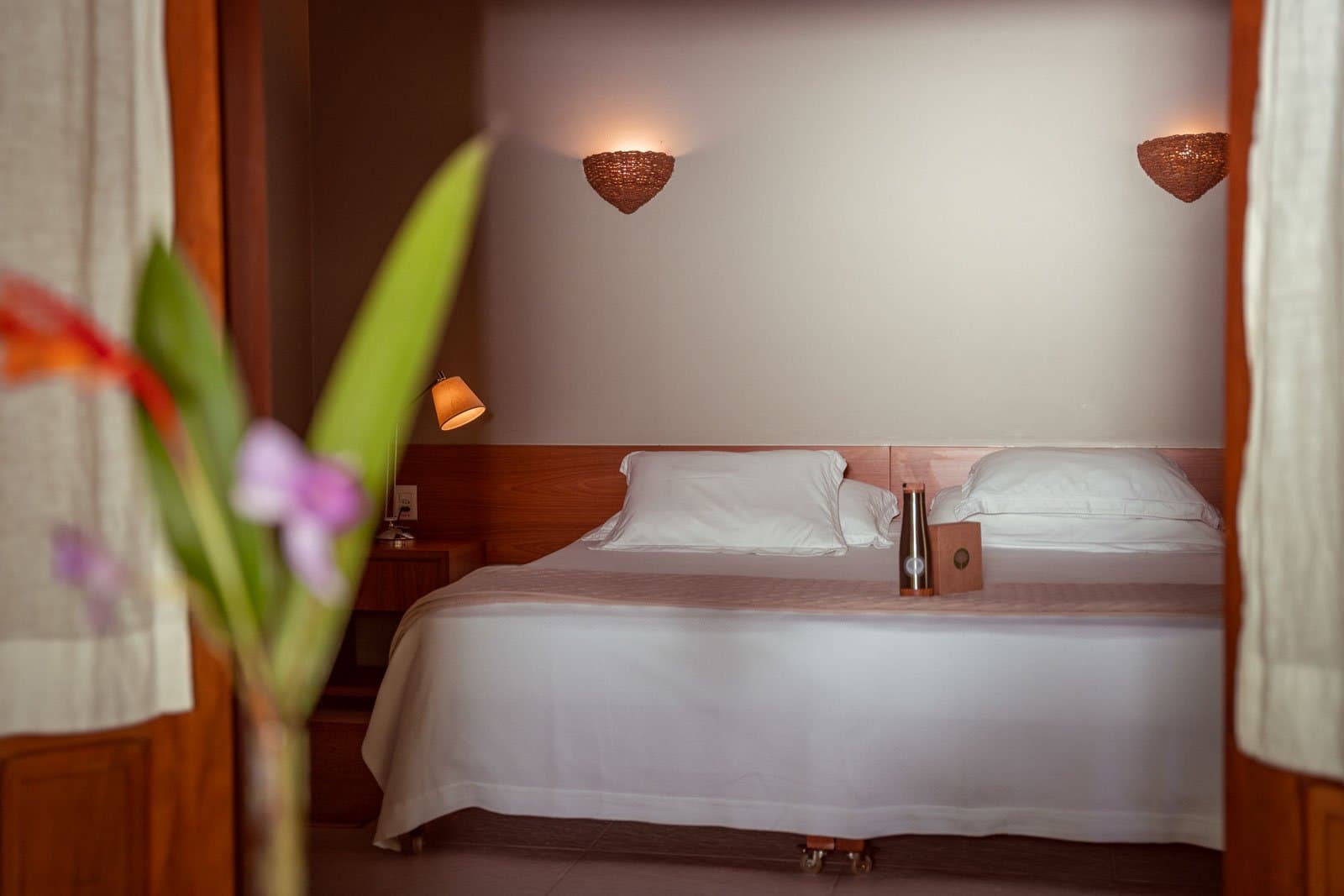 Na een dag in het regenwoud voelt dit bed als pure luxe. Een keurig opgemaakt tweepersoonsbed met strak wit linnengoed en warme houten wandlampen in een gastenkamer van Cristalino Jungle Lodge, omlijst door een wazige tropische bloem op de voorgrond