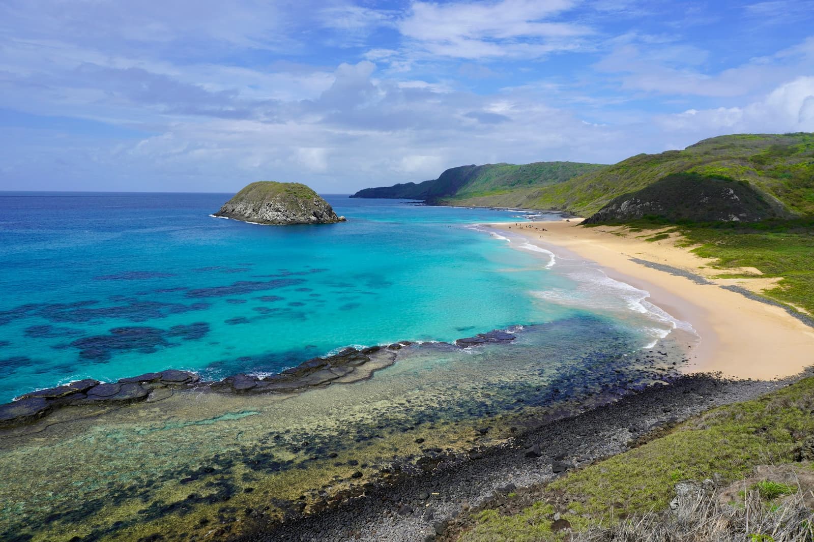 Zeewater in tinten blauw die je thuis niet kunt beschrijven. Luchtfoto van een halvemaanvormig zandstrand op Fernando de Noronha, met turquoise ondiep water dat koraalformaties onthult, vulkanische rotsplaten, een klein rotseilandje voor de kust en groene heuvels die de baai omlijsten