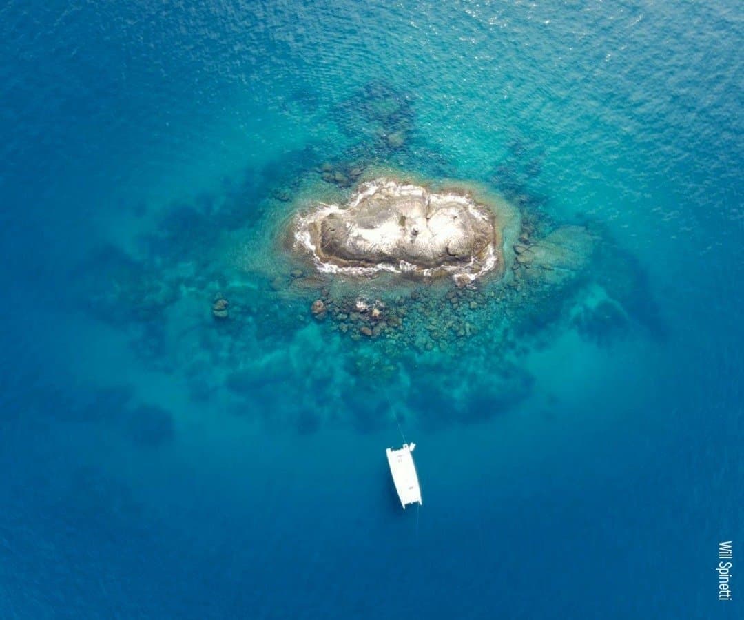 Zo'n rif wil je voor jezelf houden, en hier lukt dat nog. Luchtfoto van een witte catamaran voor anker naast een rotsachtig eilandje omgeven door kristalhelder turquoise water tijdens een duikreis naar Fernando de Noronha