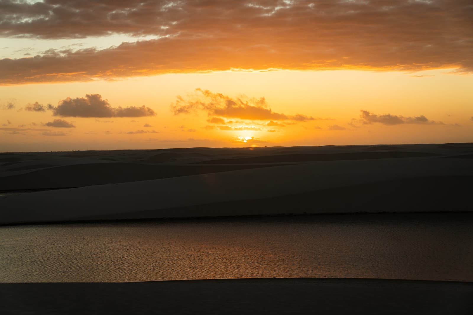 Le soleil disparaît lentement, et les dunes gardent sa couleur encore un moment. Coucher de soleil doré sur des dunes de sable en silhouette et un lagon tranquille reflétant une lumière ambrée lors du circuit à cheval dans les Lençóis Maranhenses