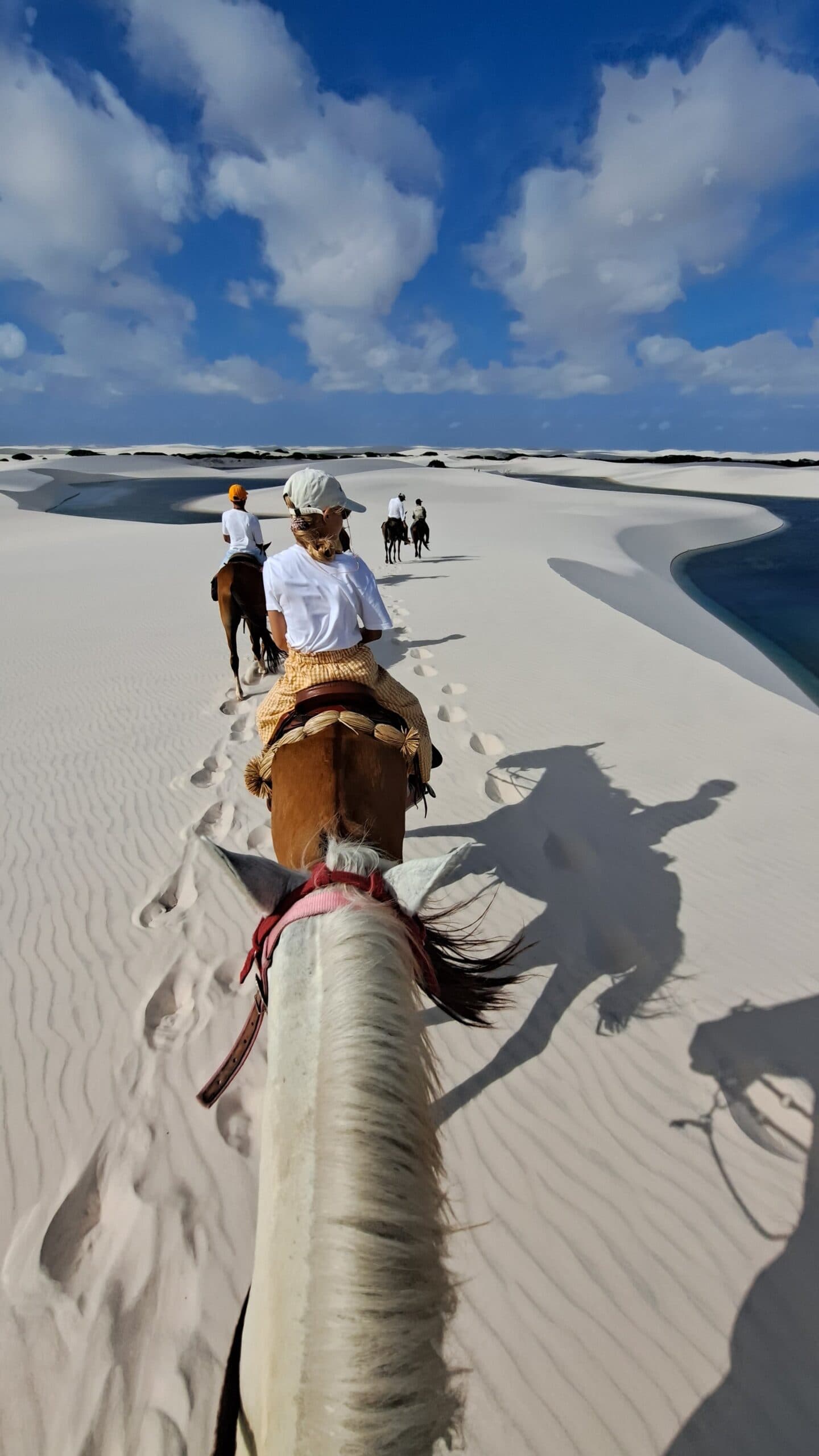 Des empreintes de sabots dans le sable blanc, une lagune à portée de regard. Le bon rythme pour découvrir les Lençóis. Vue à la première personne depuis le dos d'un cheval lors d'un tour à cheval dans les Lençóis Maranhenses, avec des cavaliers et des empreintes de sabots traversant des dunes blanches ondulées au bord d'un lagon turquoise