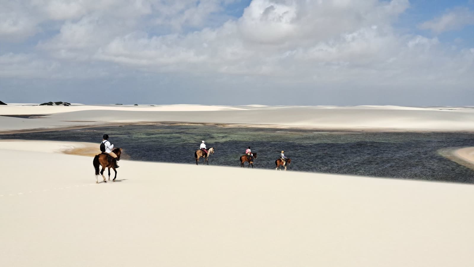 Quand le sable est votre seule boussole, un cheval suffit. Quatre cavaliers traversent des dunes de sable blanc longeant une lagune d'eau douce sombre lors d'une randonnée à cheval dans les Lençóis Maranhenses, sous un ciel partiellement nuageux