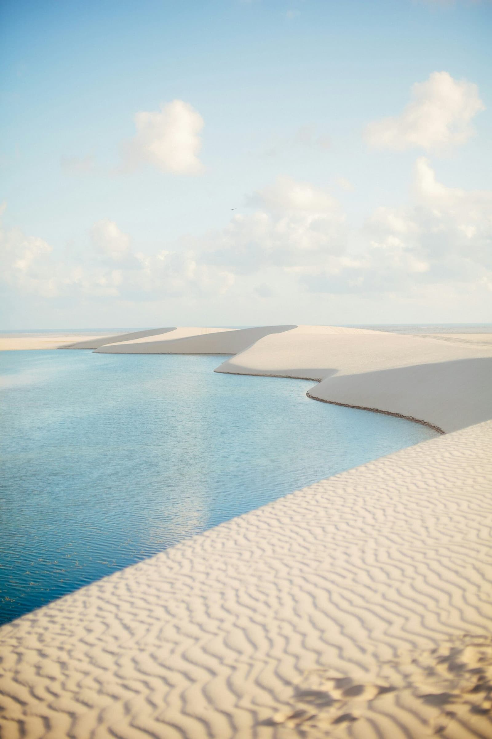Le sable et l'eau se disputent le terrain, sans jamais se départager. Lagune turquoise aux eaux cristallines encadrée par de grandes dunes de sable blanc sous un ciel bleu pâle dans le Lençóis Maranhenses