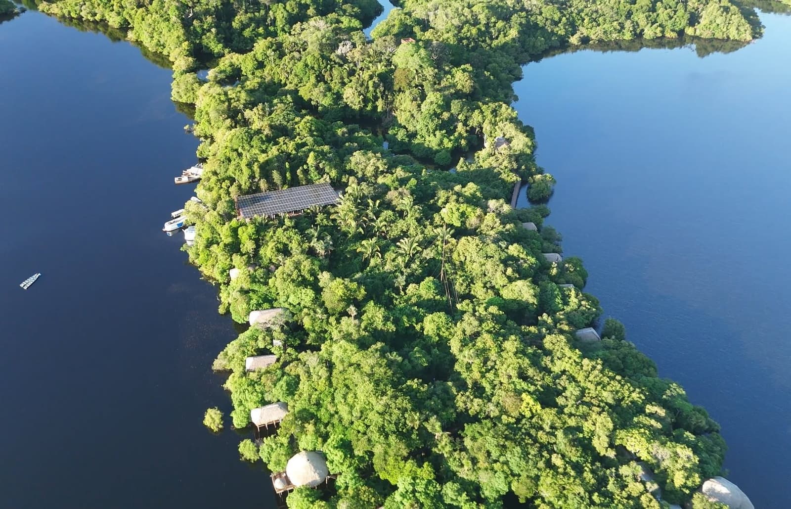 A floresta engole o lodge quase por inteiro, e isso é exatamente a ideia. Vista aérea do Juma Amazon Jungle Lodge encravado em uma ilha de densa floresta amazônica cercada por águas escuras, com bangalôs de palha, painéis solares e barcos visíveis entre a copa das árvores