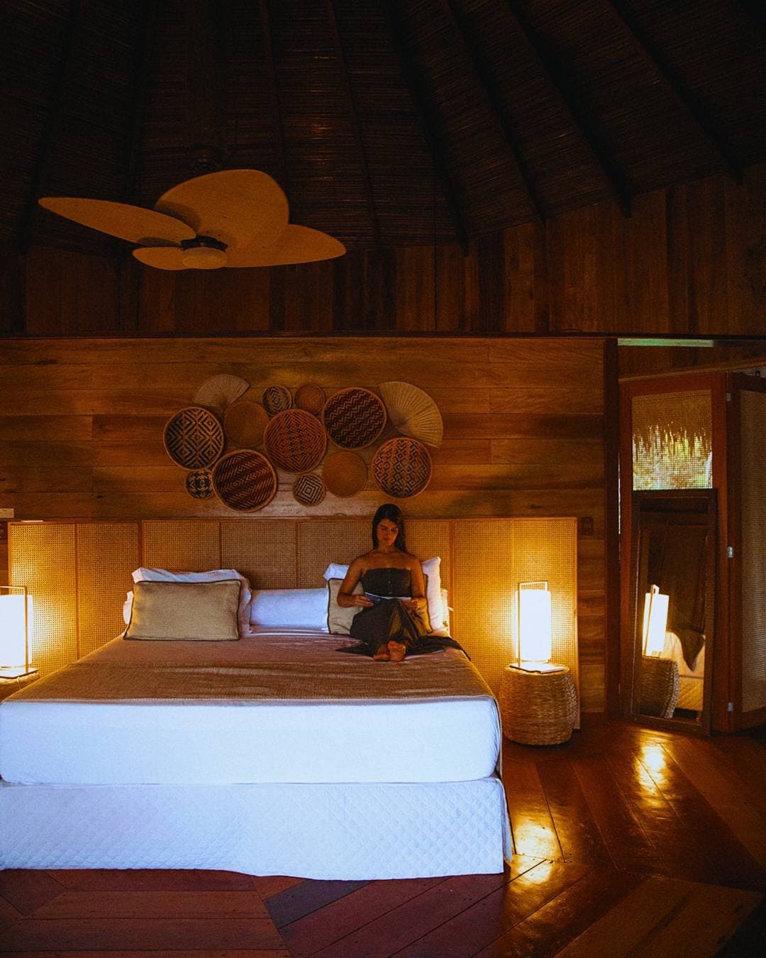 Noites na Amazônia pedem exatamente isso: silêncio, luz morna e uma boa leitura. Uma mulher lê numa cama king-size dentro de um bangalô do Juma Amazon Jungle Lodge, envolta por luz suave de abajures, cabeceira de rattan trançado, cestos decorativos nas paredes de madeira e teto de palha com ventilador de madeira