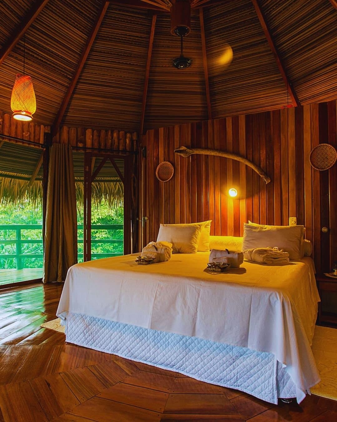 A floresta não fica do lado de fora, ela entra junto. Interior aconchegante de bungalow na Juma Amazon Jungle Lodge com cama de casal em roupa branca, teto cônico de palha, piso de madeira nobre polida e porta de varanda aberta emoldurando vegetação exuberante da floresta