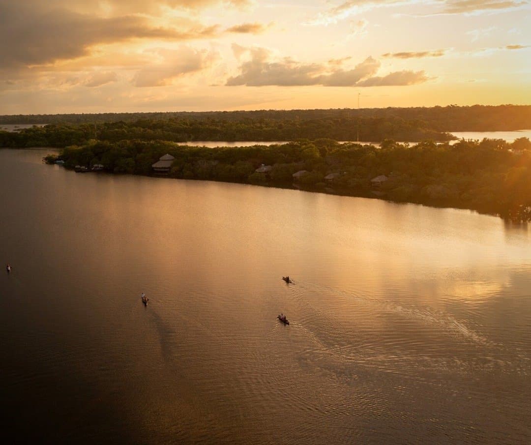Na Amazônia, o pôr do sol é convite, não enfeite. Vista aérea de pequenas canoas deslizando por um rio largo ao pôr do sol dourado, com os bangalôs de palha do Juma Amazon Jungle Lodge aninhados na densa floresta amazônica na margem oposta