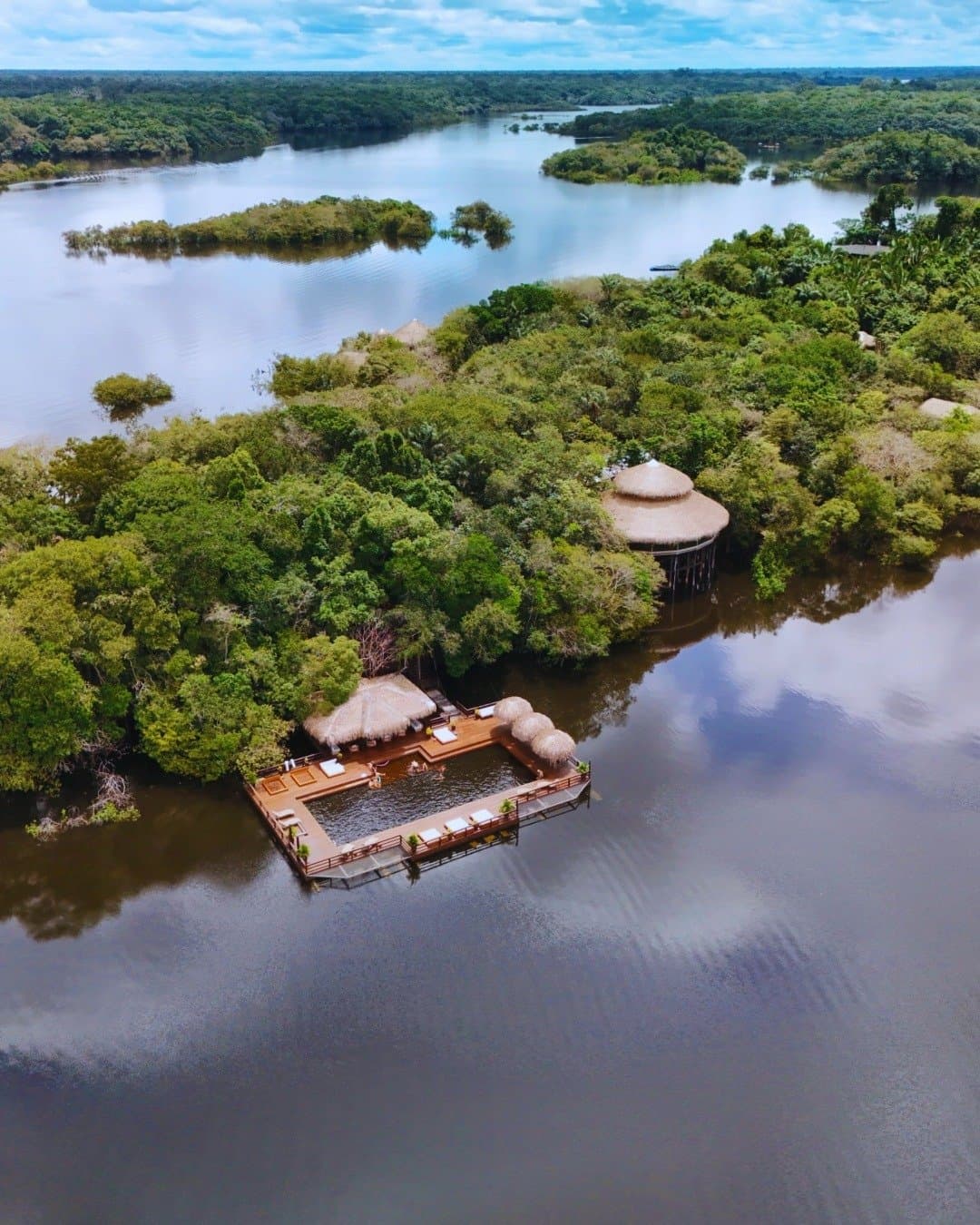 O rio ao redor, a floresta logo ali acima. Vista aérea do Juma Amazon Jungle Lodge mostrando um deck flutuante de madeira com quiosques de palha e piscina fluvial cercada por floresta amazônica inundada e águas escuras