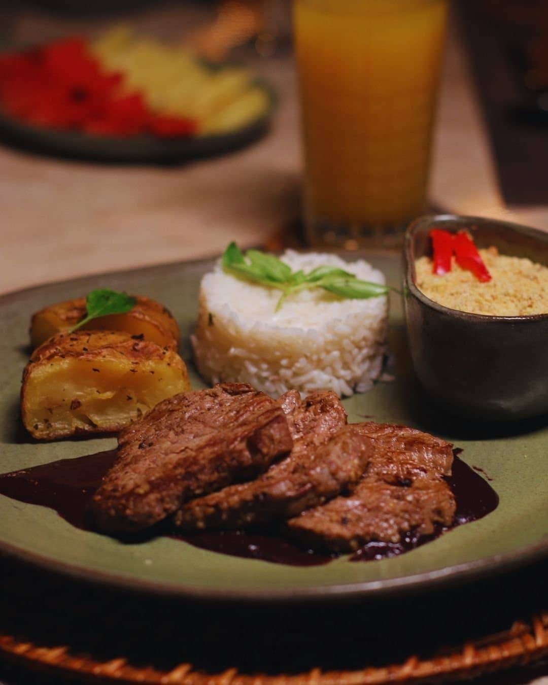 Comida de verdade, com aquele jeitão brasileiro que reconforta. Prato servido no Juma Amazon Jungle Lodge com tiras de carne bovina grelhada ao molho escuro, arroz branco moldado com ervas frescas, batatas assadas com ervas e um potinho de farofa com pimentão vermelho, acompanhados de um copo de suco de laranja