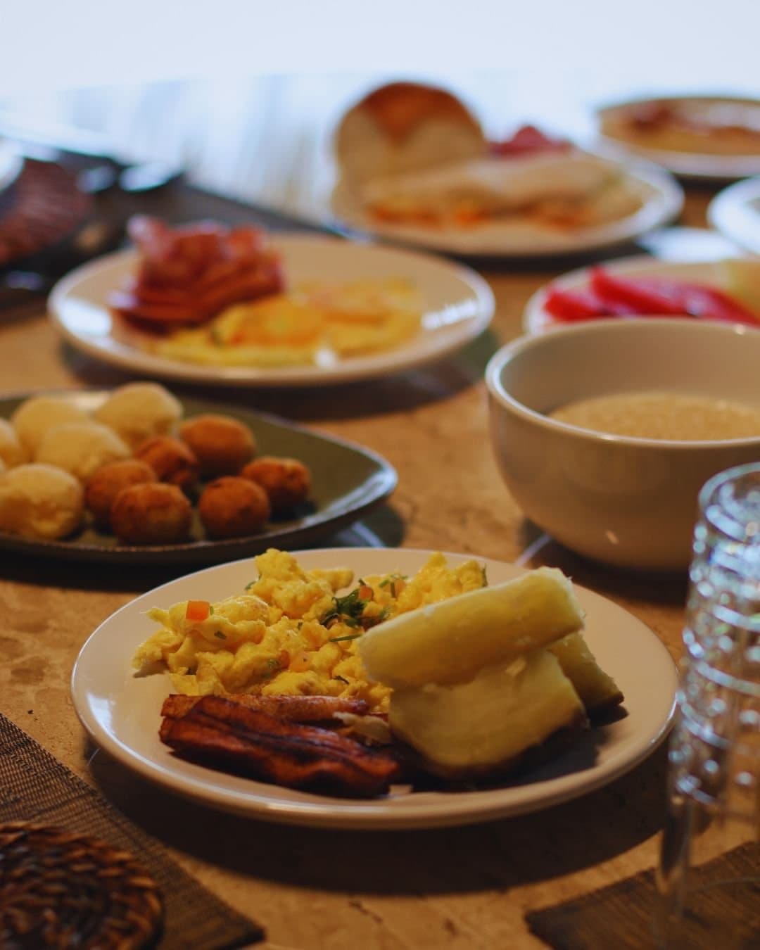 Café reforçado, porque a floresta não espera. Farto café da manhã no Juma Amazon Jungle Lodge, com ovos mexidos, bacon crocante, macaxeira cozida, frios, pimentão vermelho, salgadinhos fritos, pãezinhos e uma tigela de mingau sobre mesa de madeira