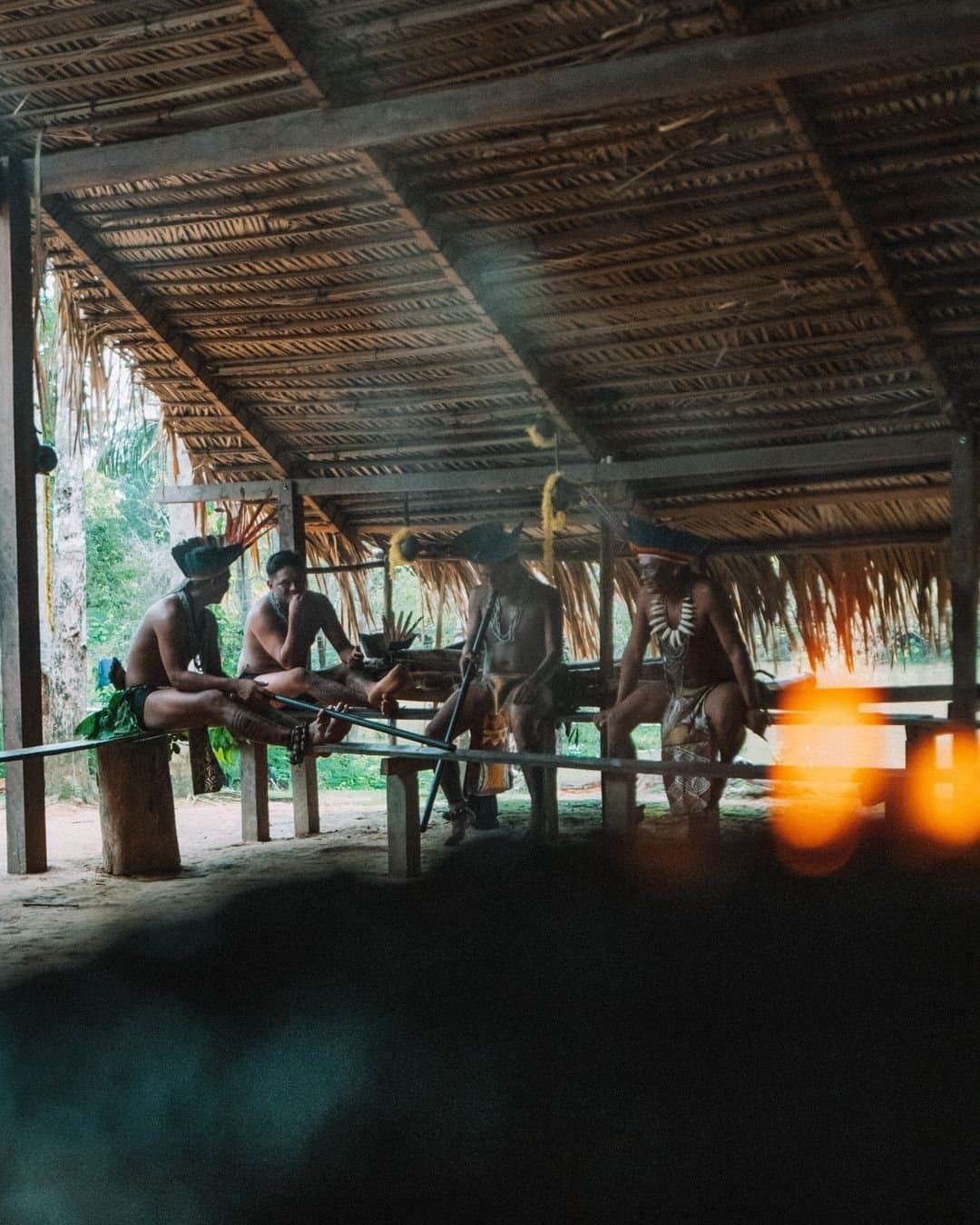 Na maloca, o tempo é dos que sabem parar para conversar. Membros de uma comunidade indígena com pintura corporal tradicional, cocares e colares de contas reunidos sob uma estrutura coberta de palha no Juma Amazon Jungle Lodge, zarabatanas compridas apoiadas em bancos de madeira