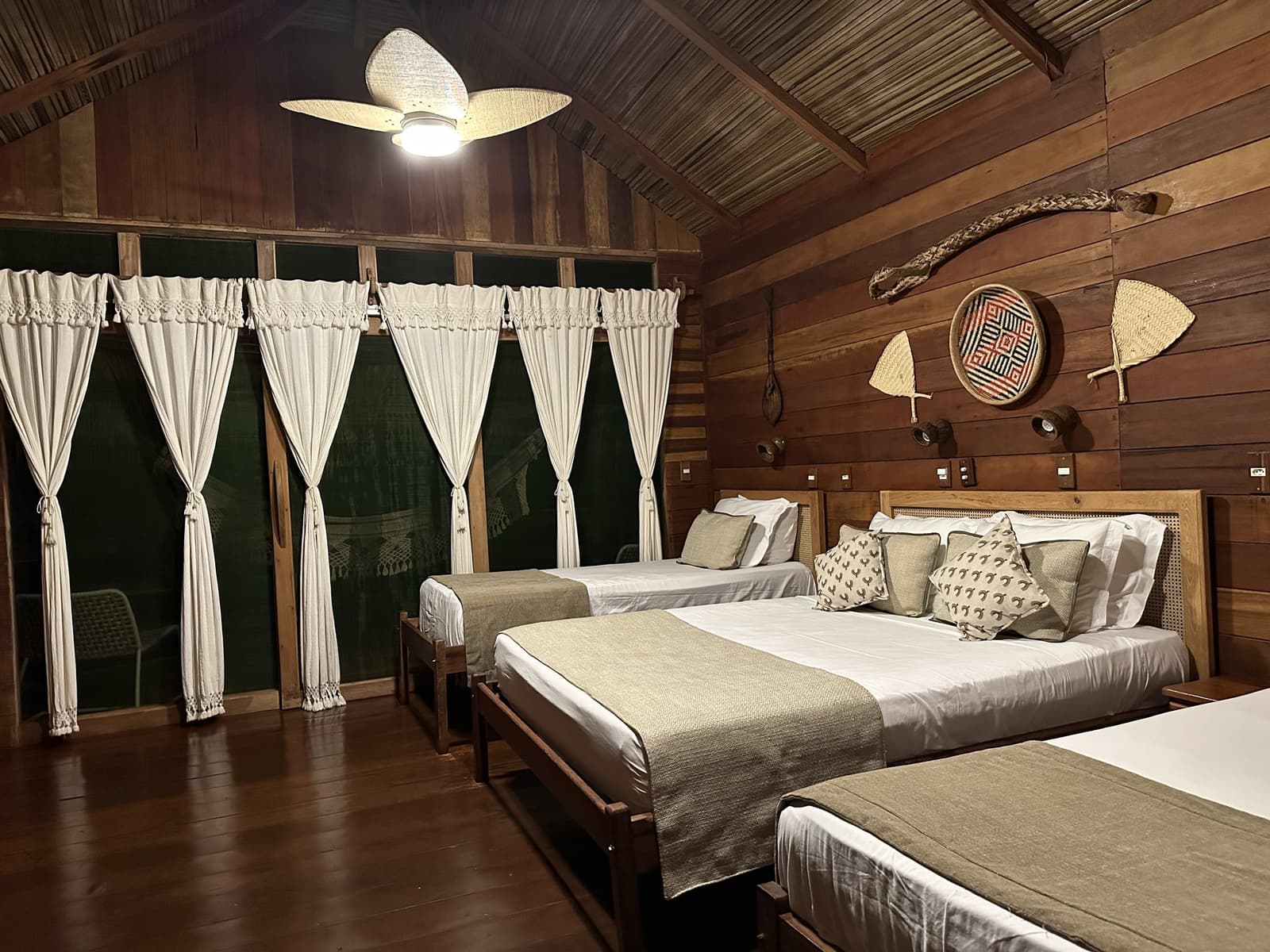 Artesanato indígena na parede, roupa de cama fresca, o conforto certo para quem dorme na floresta. Bungalow espaçoso da Juma Amazon Jungle Lodge à noite com três camas de estrutura em madeira com roupas de cama brancas, ventilador de teto trançado e artesanato indígena — cestos e remos — decorando as paredes de madeira