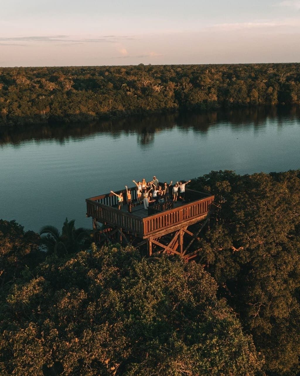 Do alto da torre, o rio e a floresta infinita. A Amazônia toda a seus pés. Um grupo de viajantes animados acena de uma alta torre de observação de madeira do Juma Amazon Jungle Lodge, rodeada por densa copa de floresta amazônica e um rio calmo ao pôr do sol