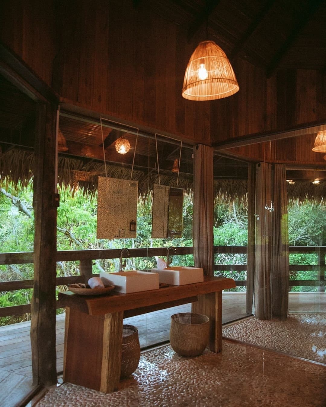 A floresta como parede, conforto que faz sentido na Amazônia. Banheiro aberto da Juma Amazon Jungle Lodge com duas cubas de apoio sobre bancada de madeira rústica, luminária trançada, piso de pedras e paredes de vidro com vista para a floresta amazônica
