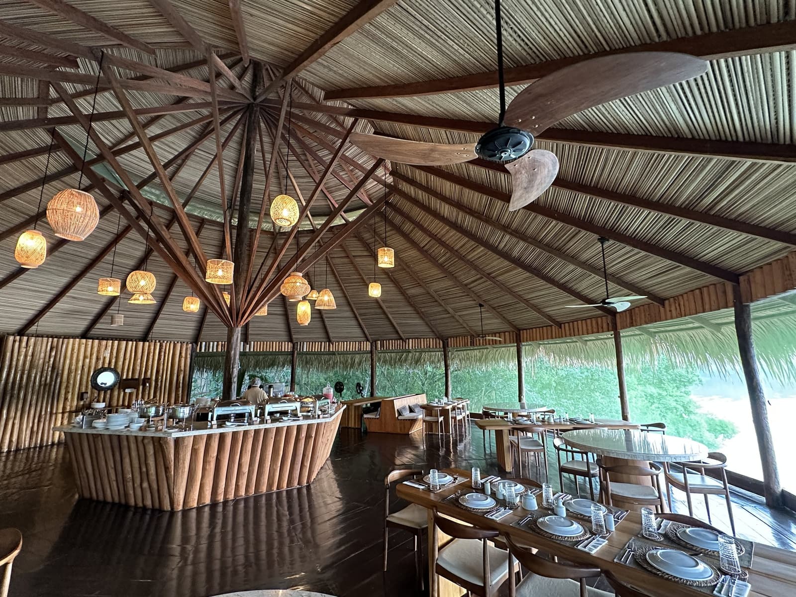A floresta fica bem pertinho enquanto você come, não tem como ignorar. O restaurante ao ar livre do Juma Amazon Jungle Lodge, com teto de palha em estrutura radial de madeira, luminárias de rattan trançado, balcão de buffet revestido de bambu e mesas de madeira postas com louça branca, abertas para a floresta exuberante