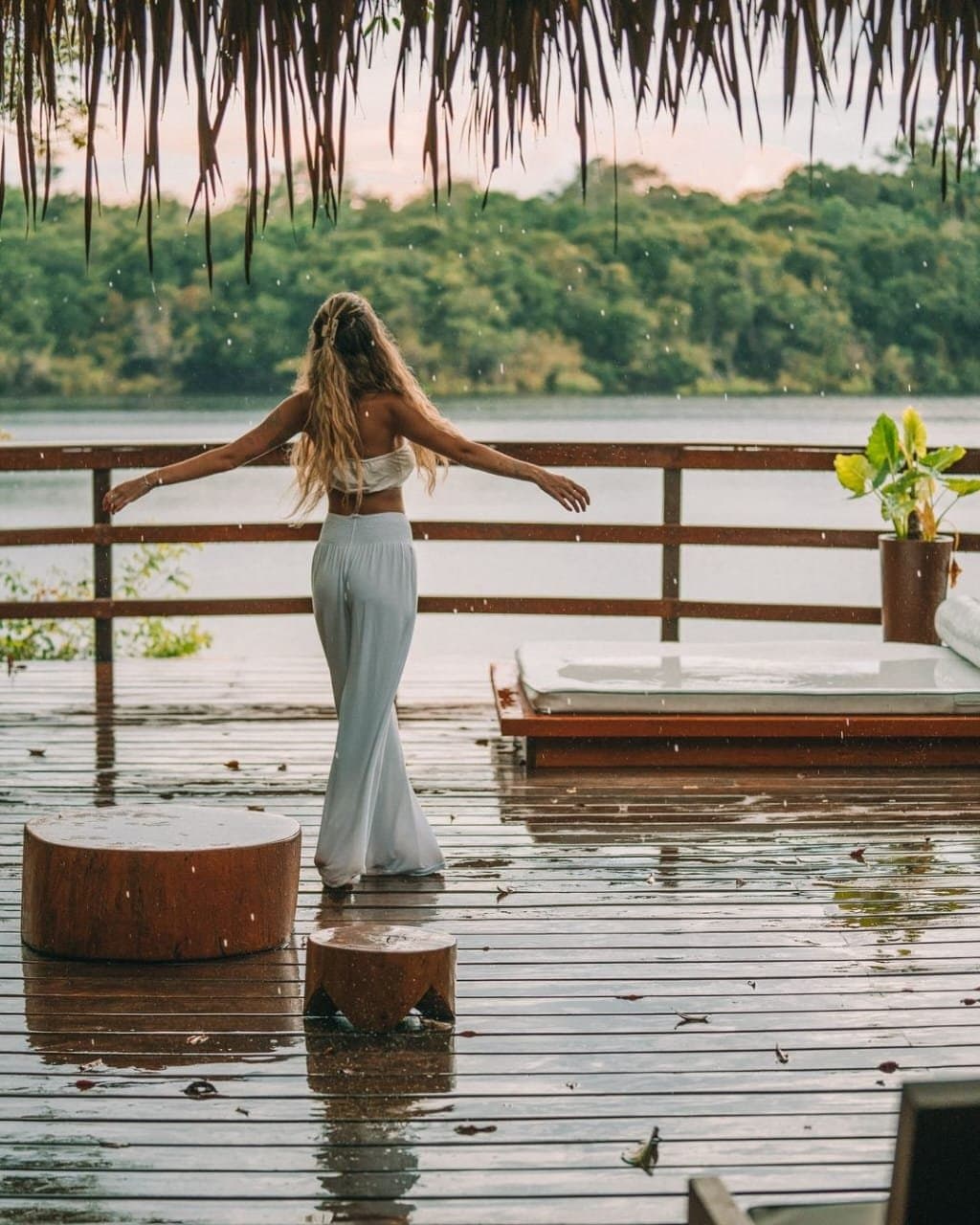 Chuva morna, braços abertos, floresta na frente. Às vezes é só isso que a gente precisa. Uma mulher de branco está de braços abertos em um deck molhado pela chuva na Juma Amazon Jungle Lodge, com o olhar voltado para um rio calmo entre a densa mata amazônica