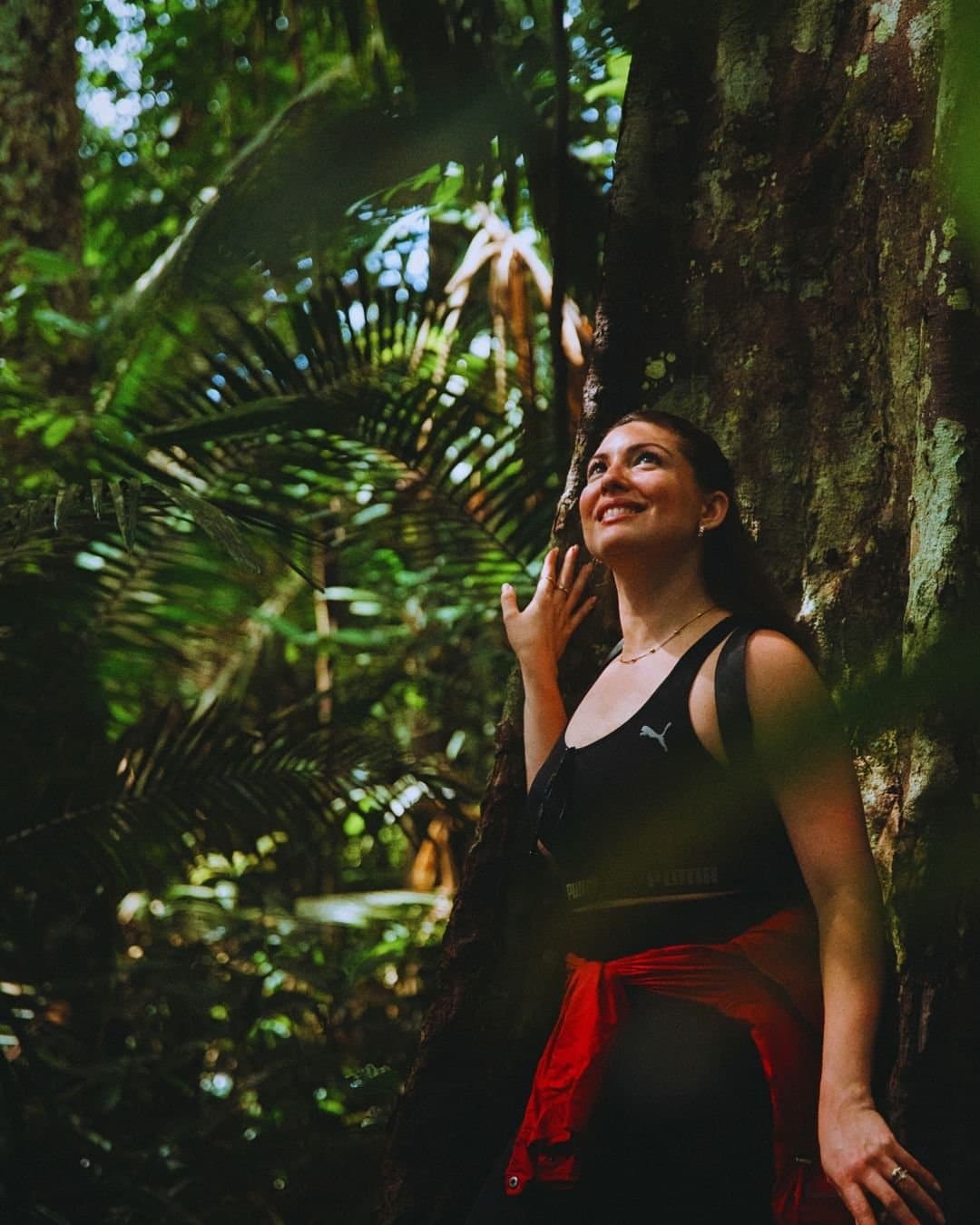 Tem árvore que faz a gente parar, respirar fundo e olhar pra cima. Uma mulher sorridente apoia-se no tronco de uma enorme árvore da floresta tropical durante uma trilha guiada do Juma Amazon Jungle Lodge, rodeada por samambaias exuberantes e palmeiras altíssimas