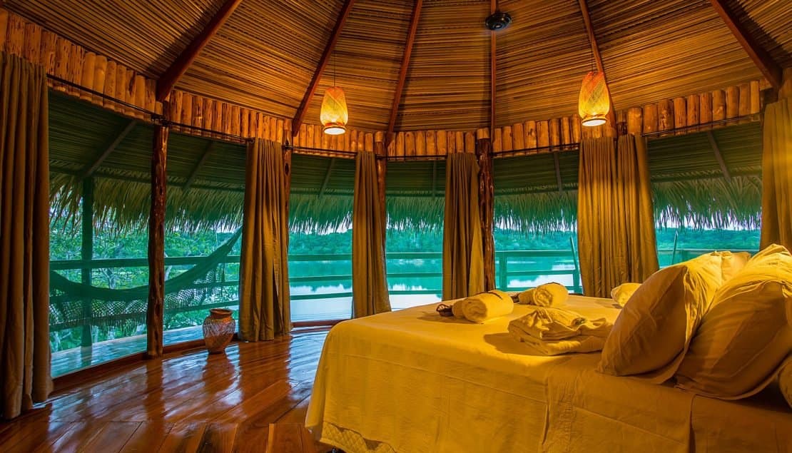 Rio como cenário, rede esperando lá fora. Isso é o que a Amazônia oferece de melhor. Interior de um bangalô do Juma Amazon Jungle Lodge com cama de casal, luminárias de palha trançada, teto de sapê e janelas abertas com vista para um rio tranquilo na floresta