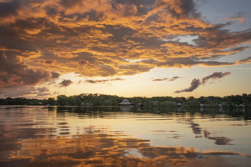 A fiery sunset sky reflected on a still river in front of the Juma Amazon Jungle Lodge, with thatched-roof bungalows nestled among dense rainforest trees along the far bank A fiery sunset sky reflected on a still river in front of the Juma Amazon Jungle Lodge, with thatched-roof bungalows nestled among dense rainforest trees along the far bank
