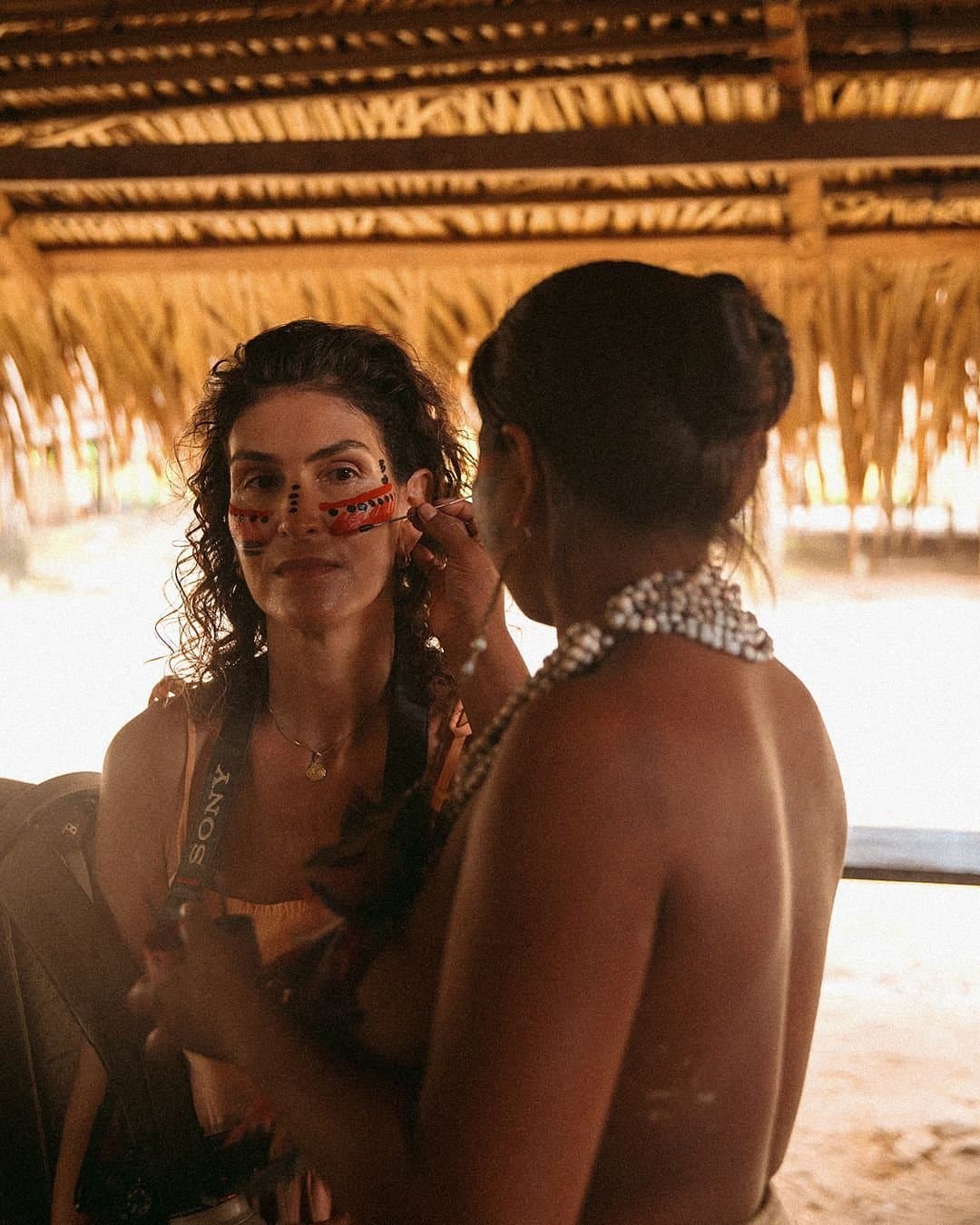 Pincel na pele, história nas mãos. Uma mulher indígena aplica pintura facial tradicional vermelha e preta em uma viajante sorridente dentro de uma oca de palha no Juma Amazon Jungle Lodge