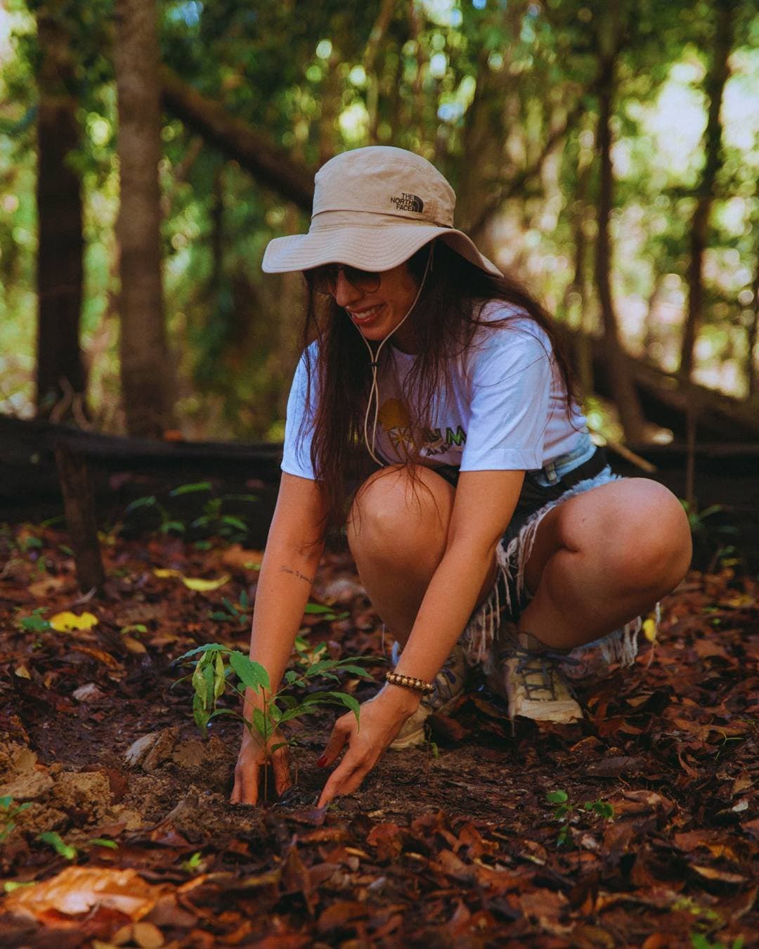 Cada muda plantada é uma promessa feita à floresta. Uma mulher sorridente de chapéu de abas largas se agacha para plantar uma muda no solo da floresta no Juma Amazon Jungle Lodge, pressionando a terra escura em torno das raízes