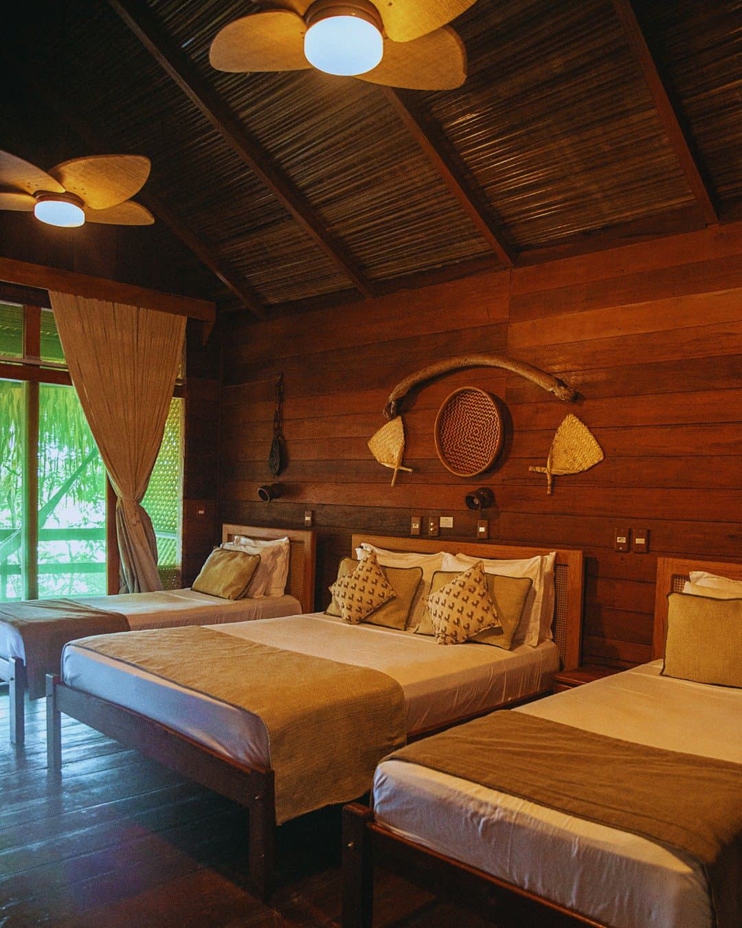 Artesanato nas paredes, floresta pela janela, um quarto que sabe onde está. Quarto triplo no Juma Amazon Jungle Lodge com três camas em madeira, decoração de parede em fibras naturais, teto de palha com ventiladores e a floresta tropical visível pela porta da varanda