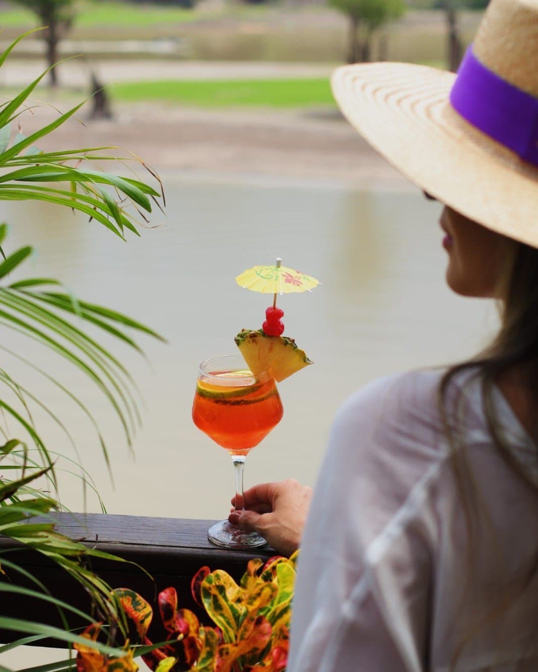 Nenhuma pressa quando o lago e a floresta estão bem ali na sua frente. Uma mulher com chapéu de palha de abas largas segura um cocktail tropical laranja vivo com abacaxi, cereja e sombrinha de papel no Juma Amazon Jungle Lodge, tendo ao fundo um curso d'água tranquilo e vegetação exuberante