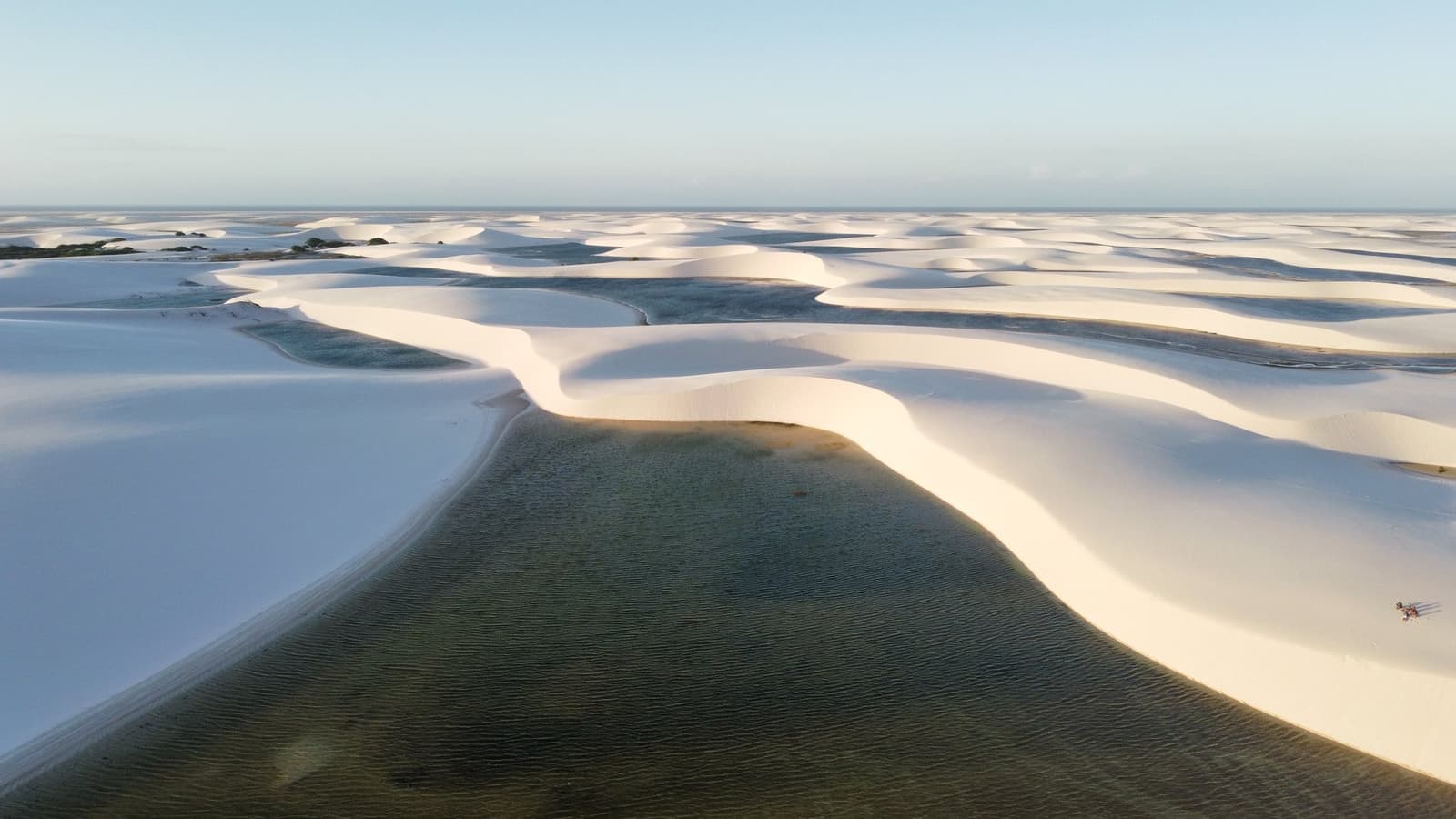 Des dunes à n'en plus finir, un lagon vert niché entre les crêtes — le parc dans toute sa démesure. Vue aérienne du trek dans les Lençóis Maranhenses, avec d'immenses dunes de sable blanc encadrant un lagon d'eau douce vert foncé à perte de vue sous un ciel bleu pâle