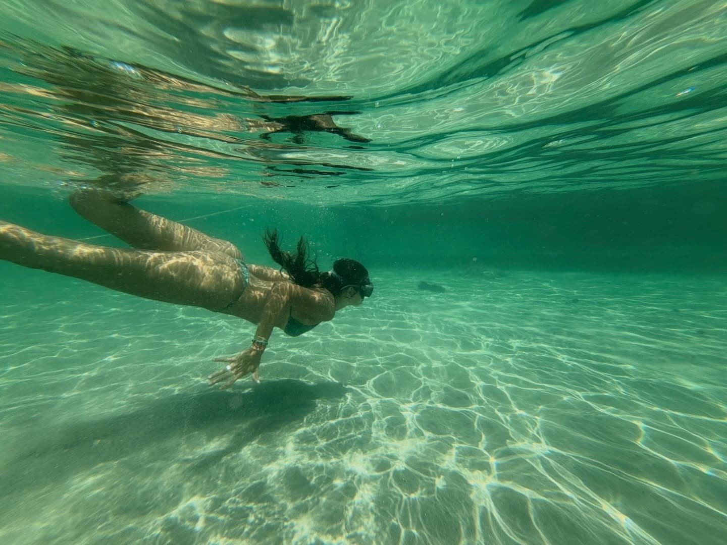 Eau émeraude, lumière ondulante — chaque lagon est une promesse de fraîcheur. Une femme plonge en apnée dans une eau turquoise cristalline au-dessus d'un fond sableux, photographiée sous l'eau lors d'une randonnée dans les Lençóis Maranhenses