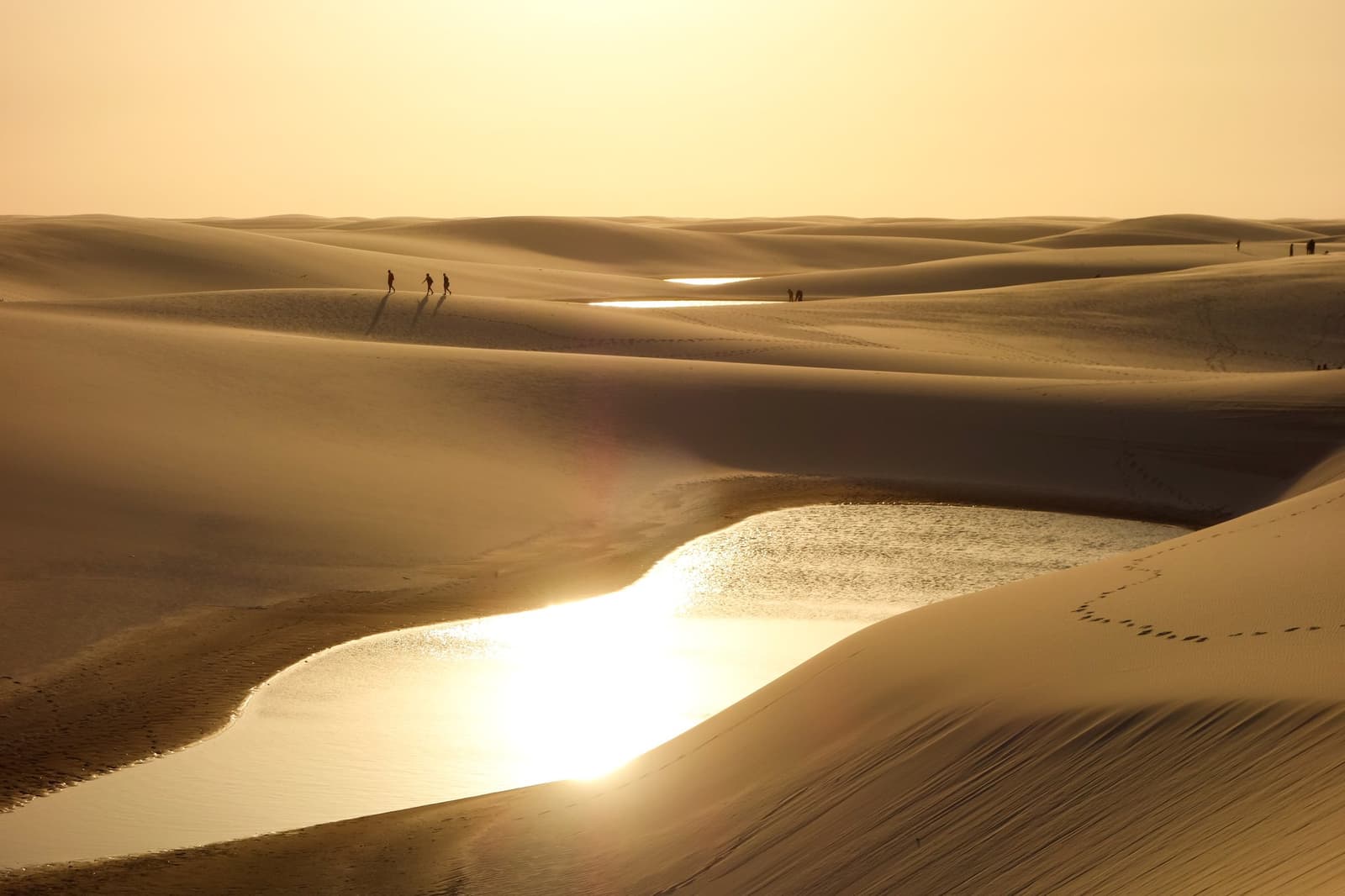 La lumière dorée inonde les dunes, et les lagunes brillent comme des miroirs. Photo: caio acquesta Silhouettes de randonneurs traversant de vastes dunes de sable à l'heure dorée lors d'une randonnée dans les Lençóis Maranhenses, avec des lagunes d'eau douce brillant entre les dunes
