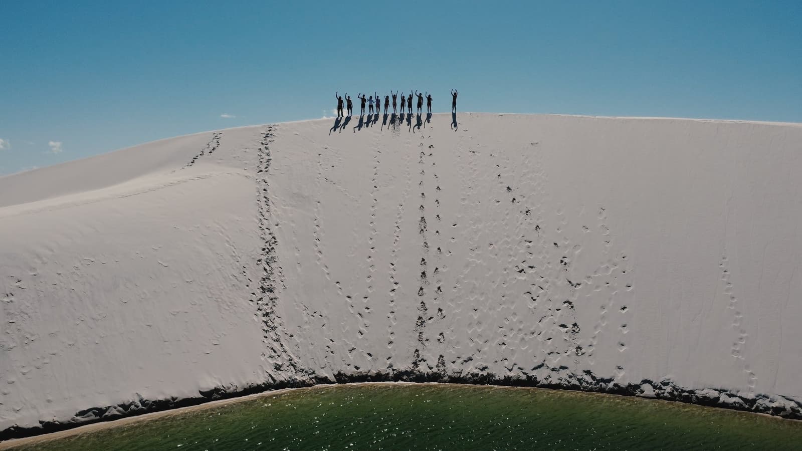Douze silhouettes, bras levés, au sommet du monde de sable. Un groupe de randonneurs en silhouette contre un ciel bleu au sommet d'une immense dune de sable blanc lors d'une randonnée dans les Lençóis Maranhenses, avec une lagune verte visible au pied de la dune