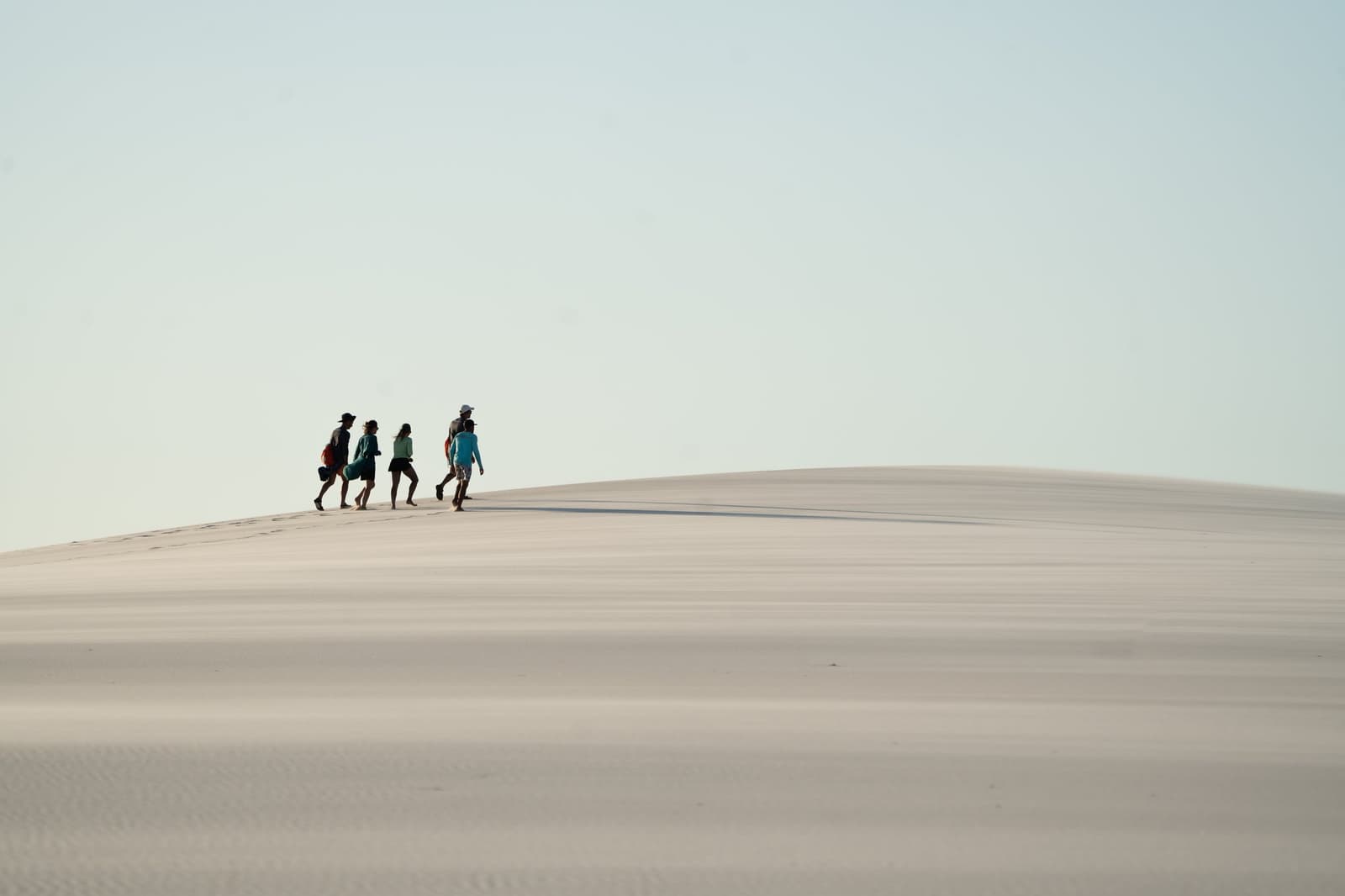 Quatre silhouettes au sommet d'une dune infinie — l'immensité des Lençóis, sans un mot. Quatre randonneurs marchant sur la crête d'une immense dune de sable blanc lors d'une randonnée dans les Lençóis Maranhenses, petites silhouettes sous un ciel pâle