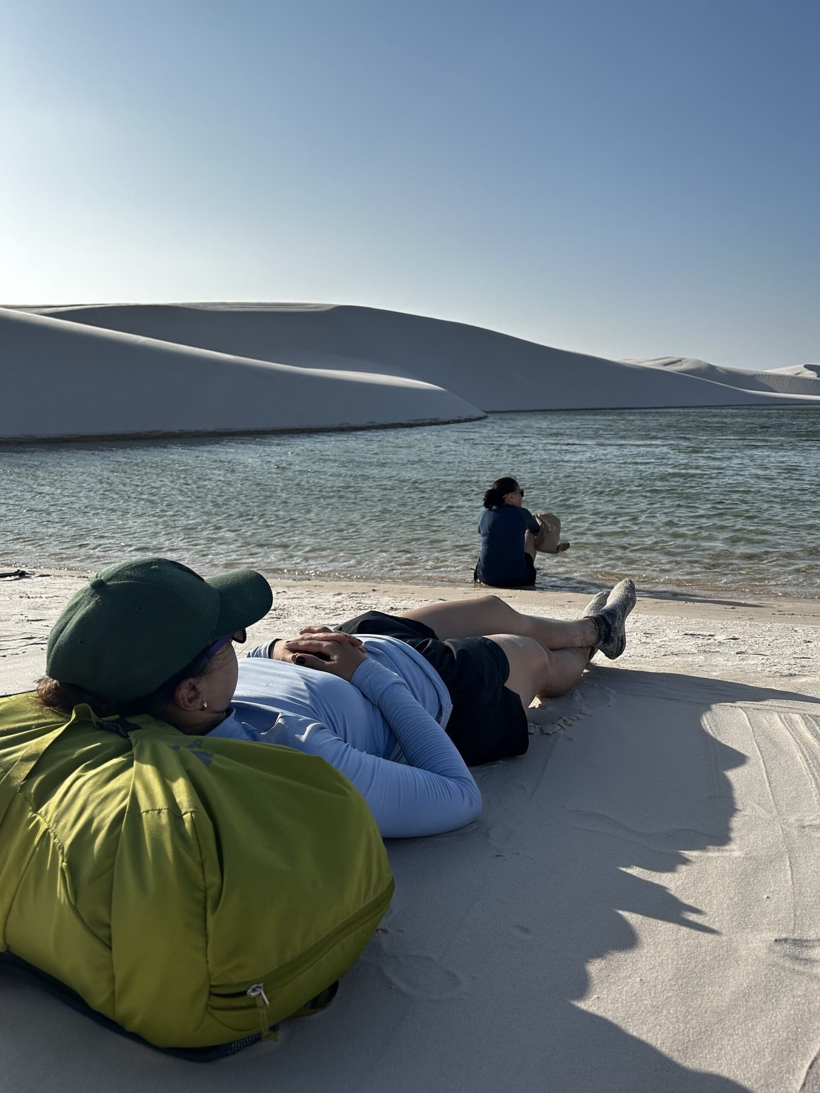 Sable blanc, eau pure — le repos après l'effort. Une randonneuse se repose sur les dunes blanches au bord d'une lagune d'eau douce lors d'une randonnée dans les Lençóis Maranhenses, une autre voyageuse assise au bord de l'eau face aux grandes dunes