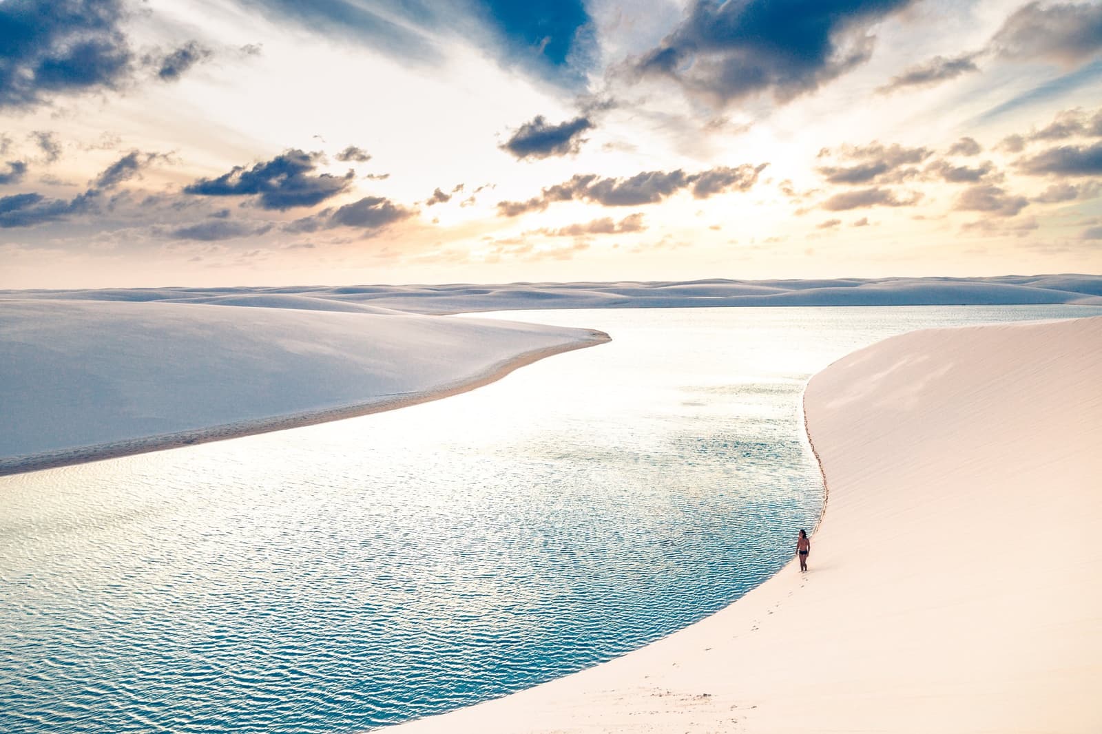 Un seul marcheur, une lagune infinie, un ciel qui s'embrase. Photo: cokada Un promeneur solitaire longe le bord d'une lagune d'eau douce cristalline entre de grandes dunes blanches lors d'une randonnée dans les Lençóis Maranhenses, sous un ciel de coucher de soleil spectaculaire