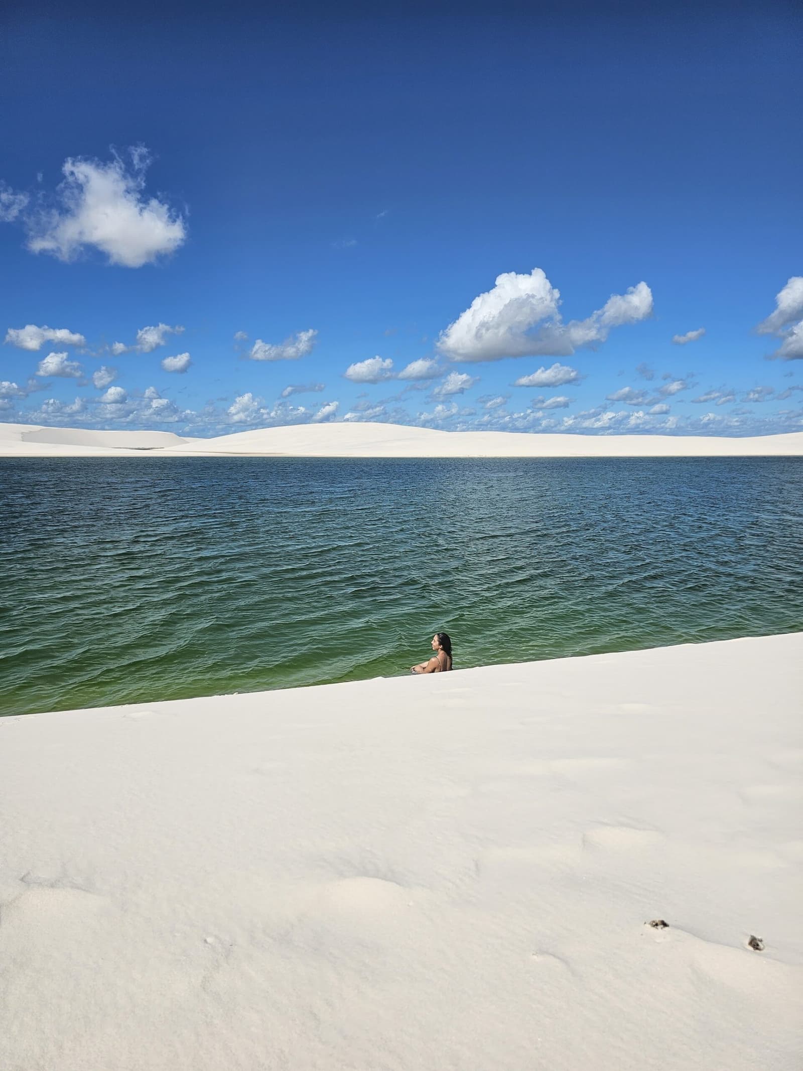 Seule dans l'émeraude, des dunes blanches à perte de vue. Une nageuse seule, à mi-corps dans une vaste lagune d'eau douce verte lors de la randonnée dans les Lençóis Maranhenses, encadrée par des dunes blanches et un ciel bleu éclatant